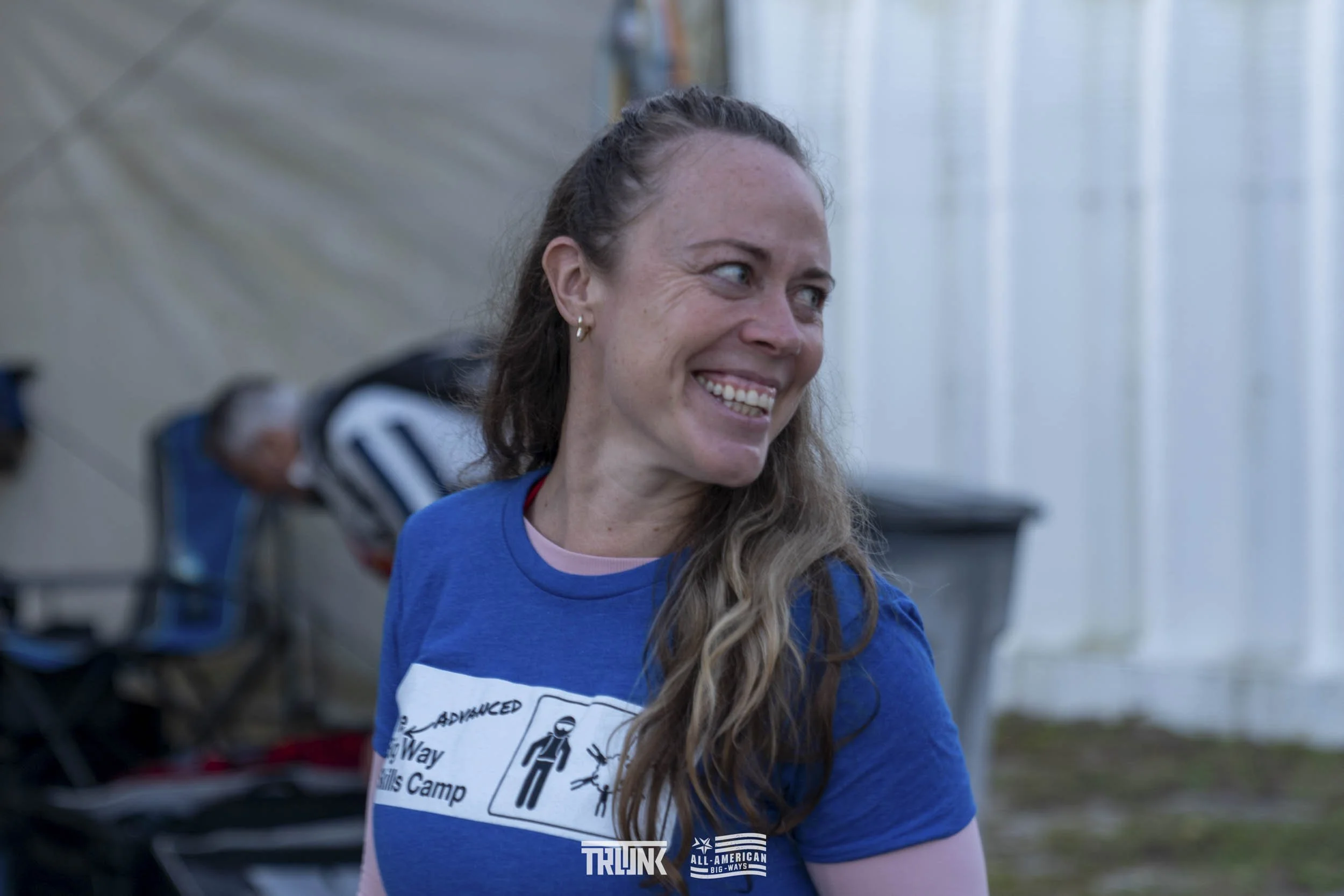 A smiling woman with long curly hair wearing a blue t-shirt with white graphics, standing outdoors near a white fence.