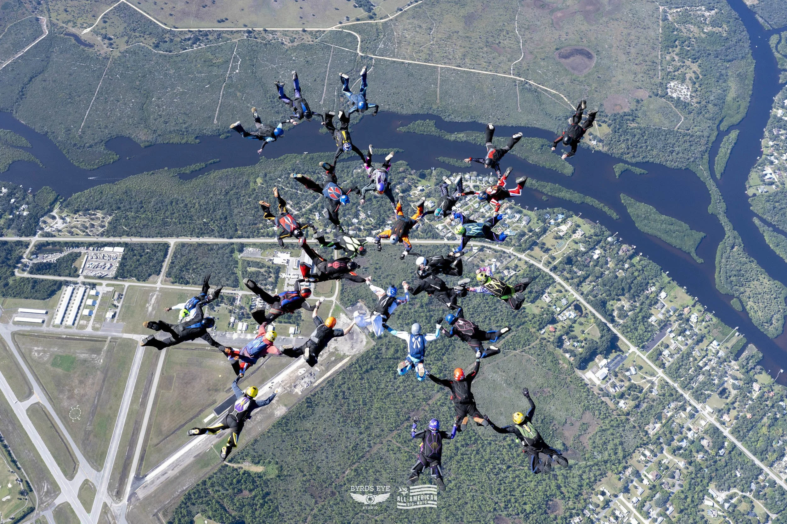 Group of skydivers floating in formation above a landscape with rivers, roads, and buildings.