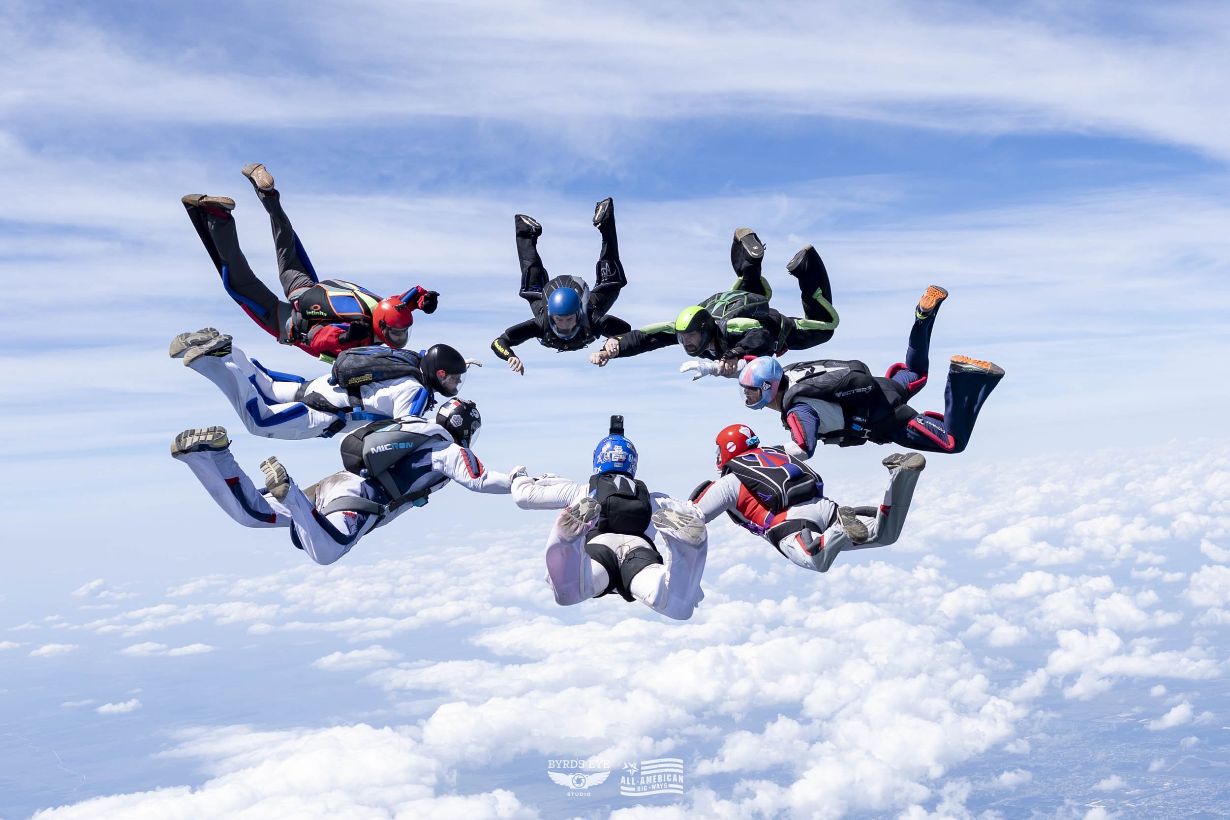Skydivers forming a circle while free-falling above clouds, wearing helmets and jumpsuits.
