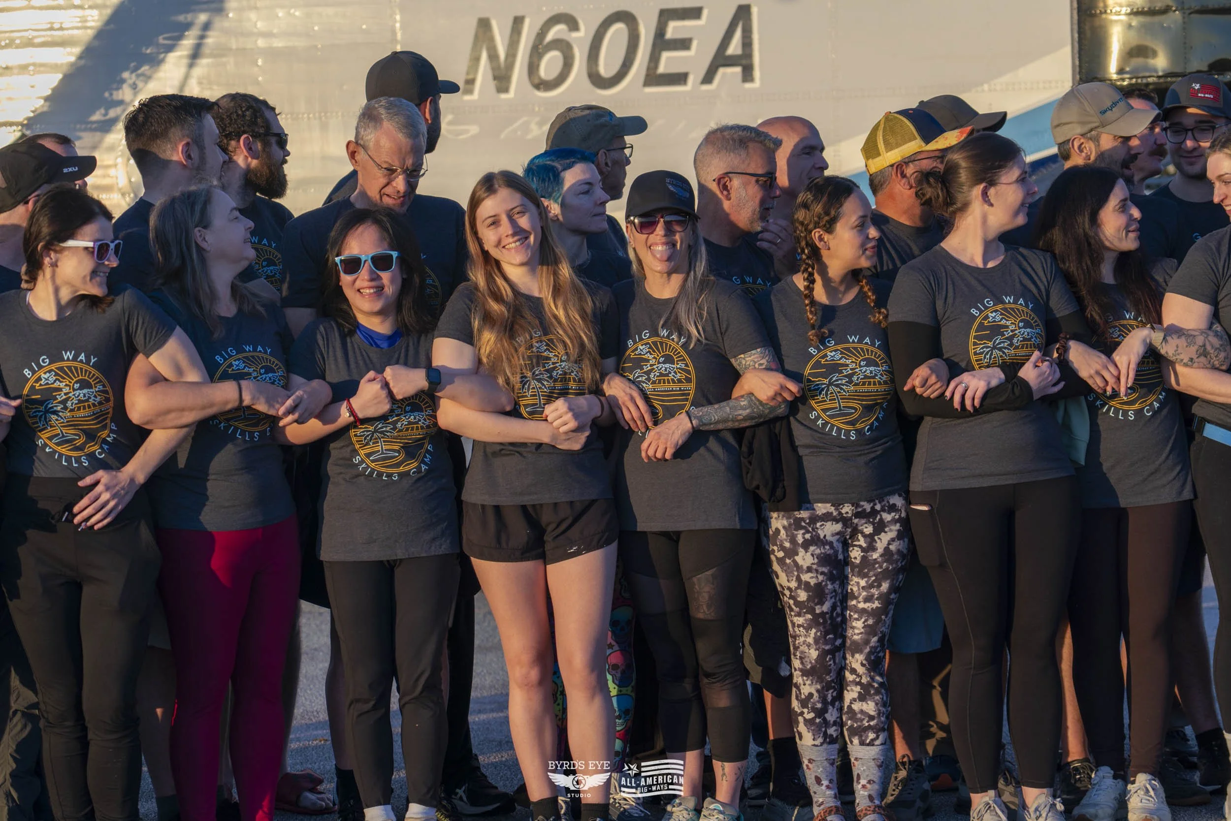Group of people in matching dark gray shirts with a colorful logo, standing in a line and holding each other's arms, outdoors with a large airplane in the background during sunset.