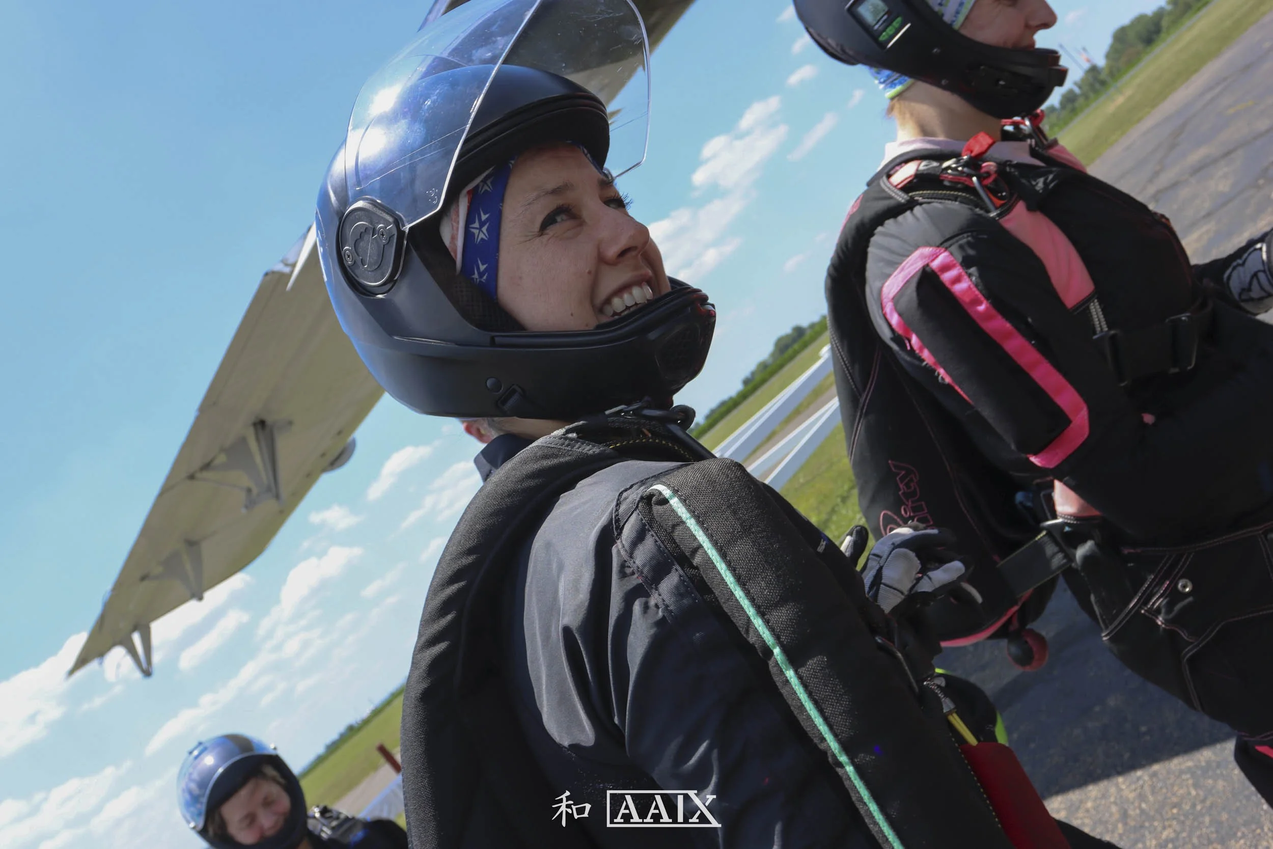 Two people wearing motorcycle gear and helmets standing outdoors on a sunny day, with a plane in the background.
