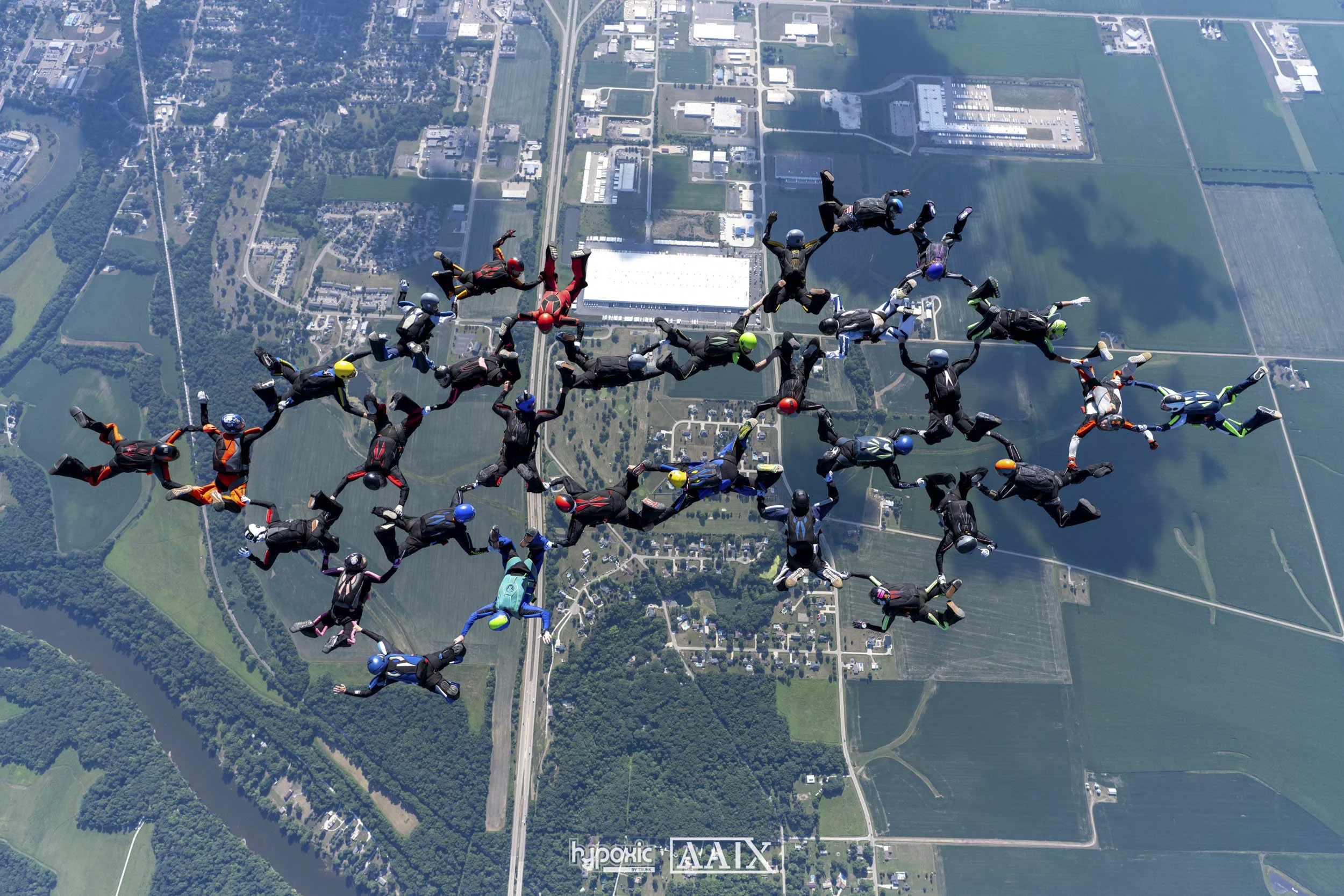 Group of skydivers forming a giant human chain in mid-air over a landscape with fields, forests, and buildings.