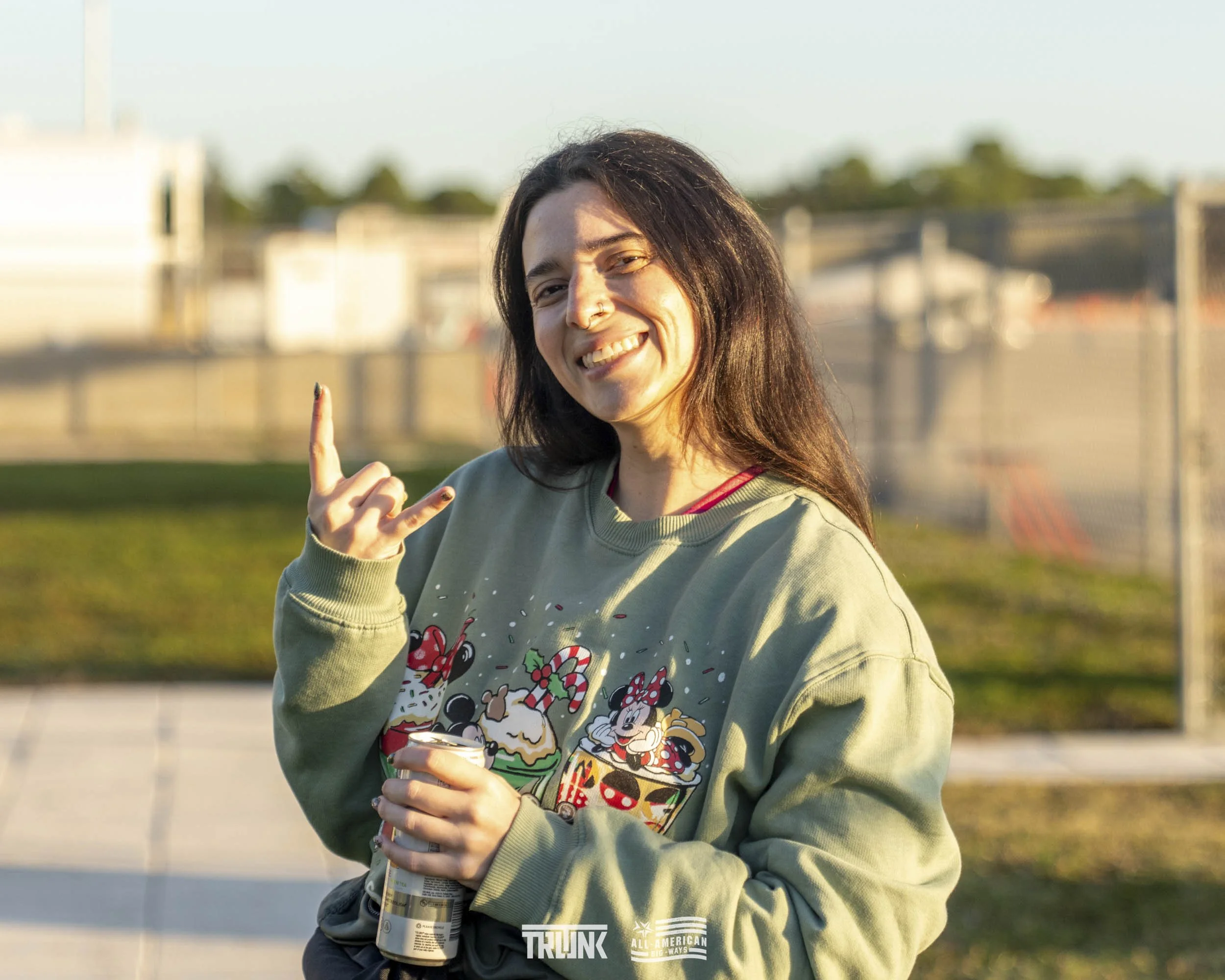 A woman with long dark hair, smiling and making a rock and roll gesture with her right hand, stands outdoors at sunset wearing a green sweatshirt with colorful Christmas-themed characters.