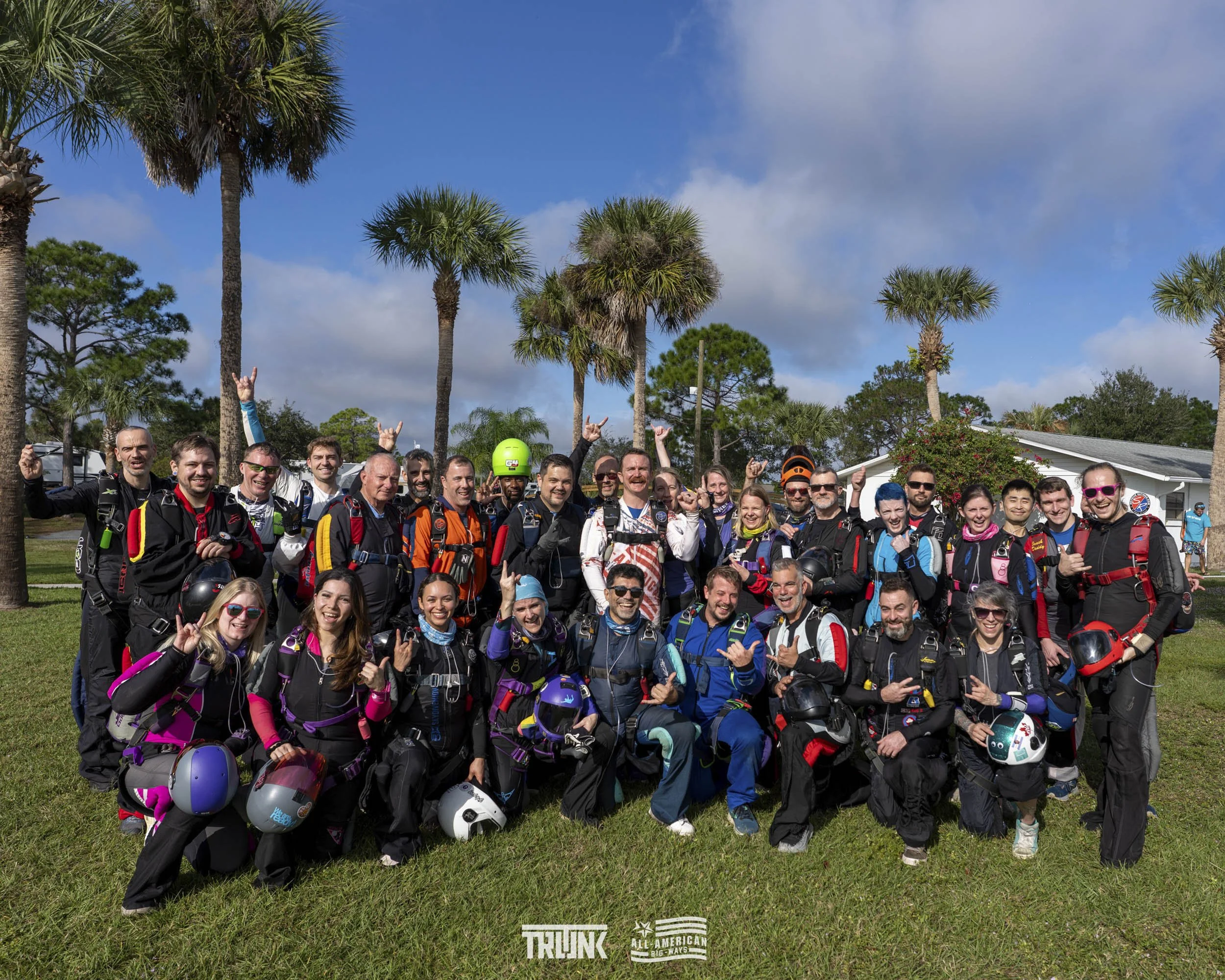 Group of skydivers in skydiving jumpsuits and helmets, posing outdoors on grass with palm trees and a building in the background, celebrating after a skydiving jump.