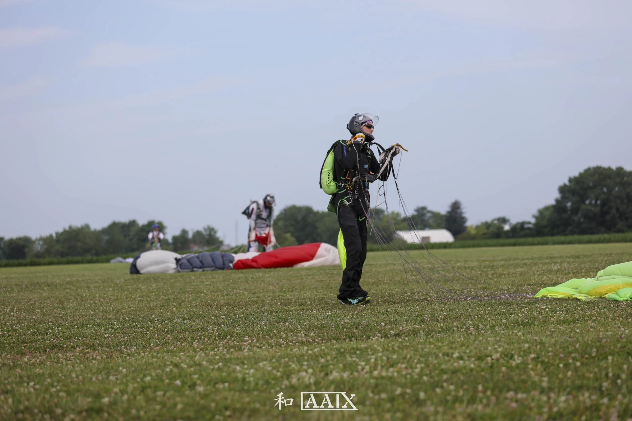 A person dressed in skydiving gear holding parachute lines on a grassy field with other skydivers and parachutes in the background.