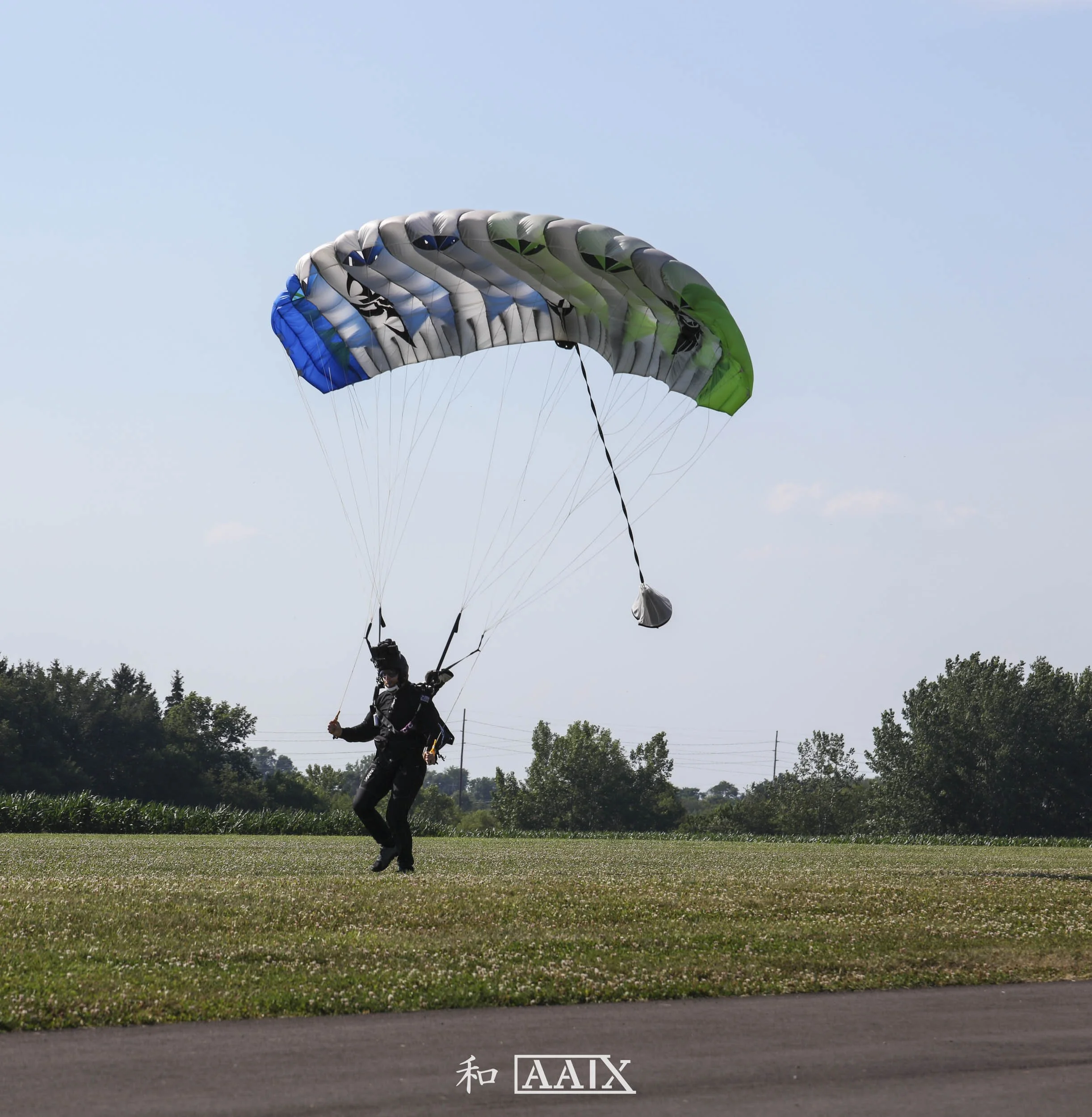 Person preparing for skydiving with a parachute on a grassy field near trees and power lines under a blue sky.
