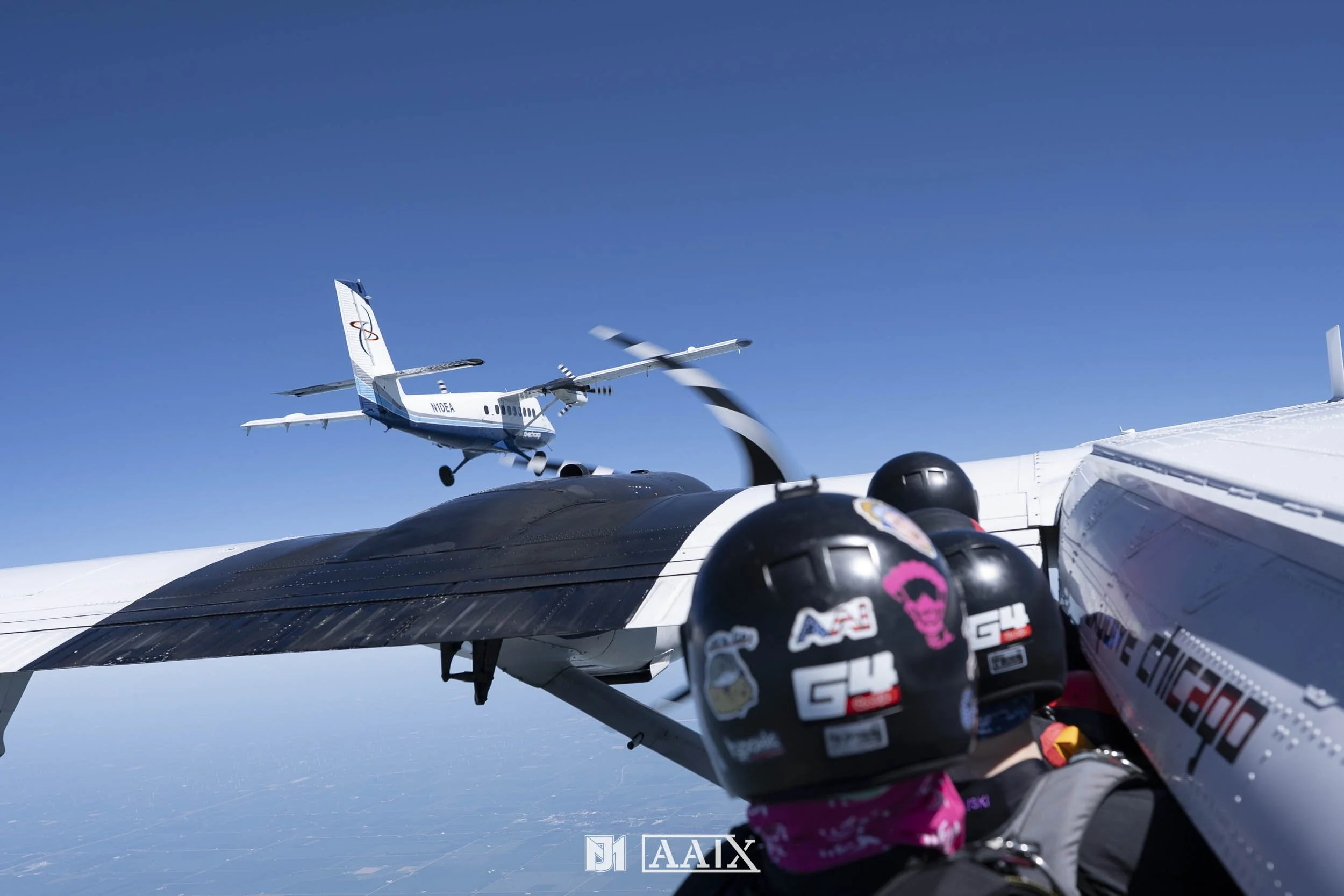 A person in a helmet and jumpsuit taking a selfie inside a cockpit, with a black aircraft wing visible, while a propeller airplane is flying in the clear blue sky in the background.