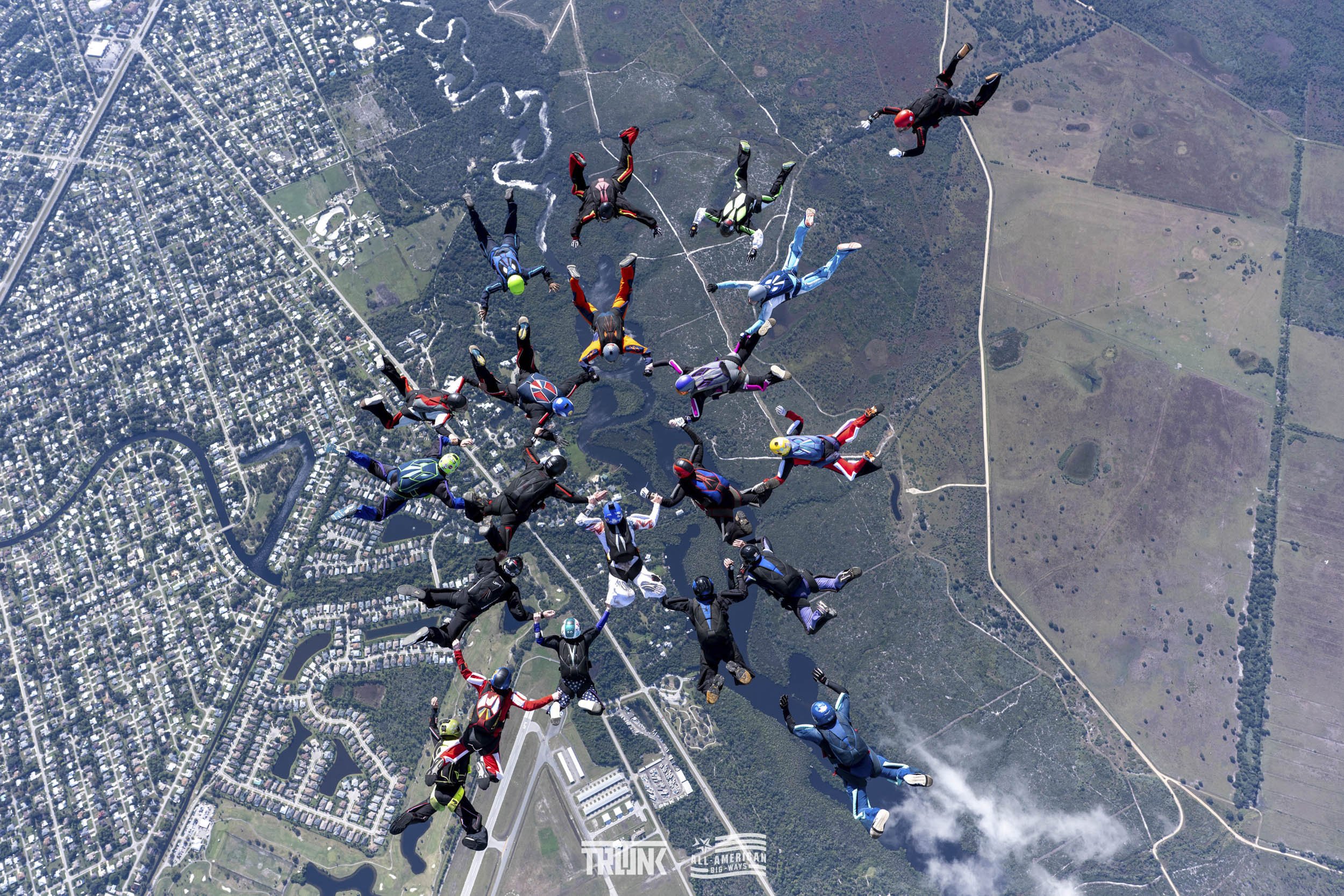 Group of skydivers in freefall over a suburban and rural landscape, holding hands in a circle, with some wearing colorful jumpsuits and helmets.