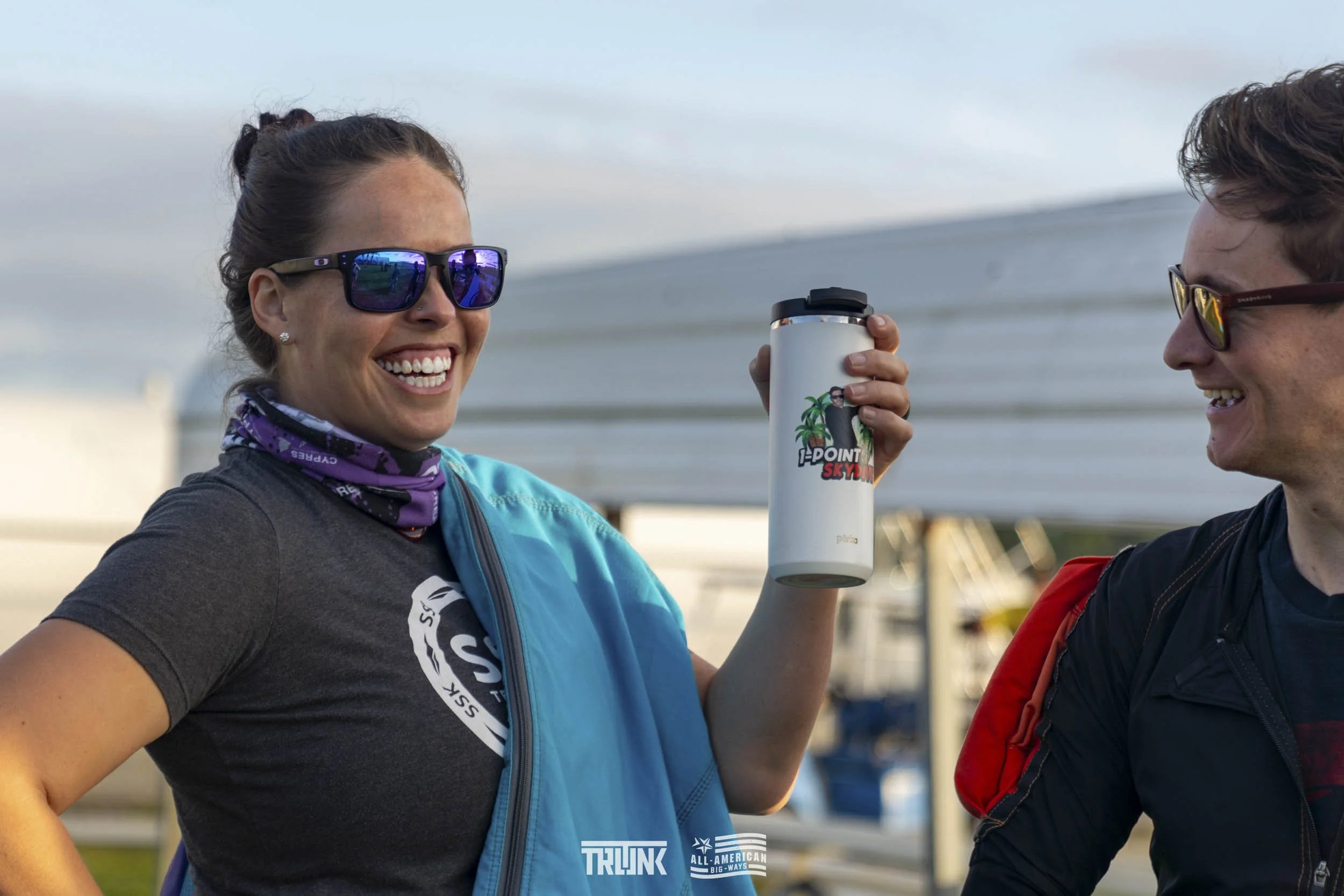 Two smiling people wearing sunglasses, standing outdoors near boats, with one holding a white travel mug that has a logo of a tropical scene and the text 'H-Point Sky Dive'.