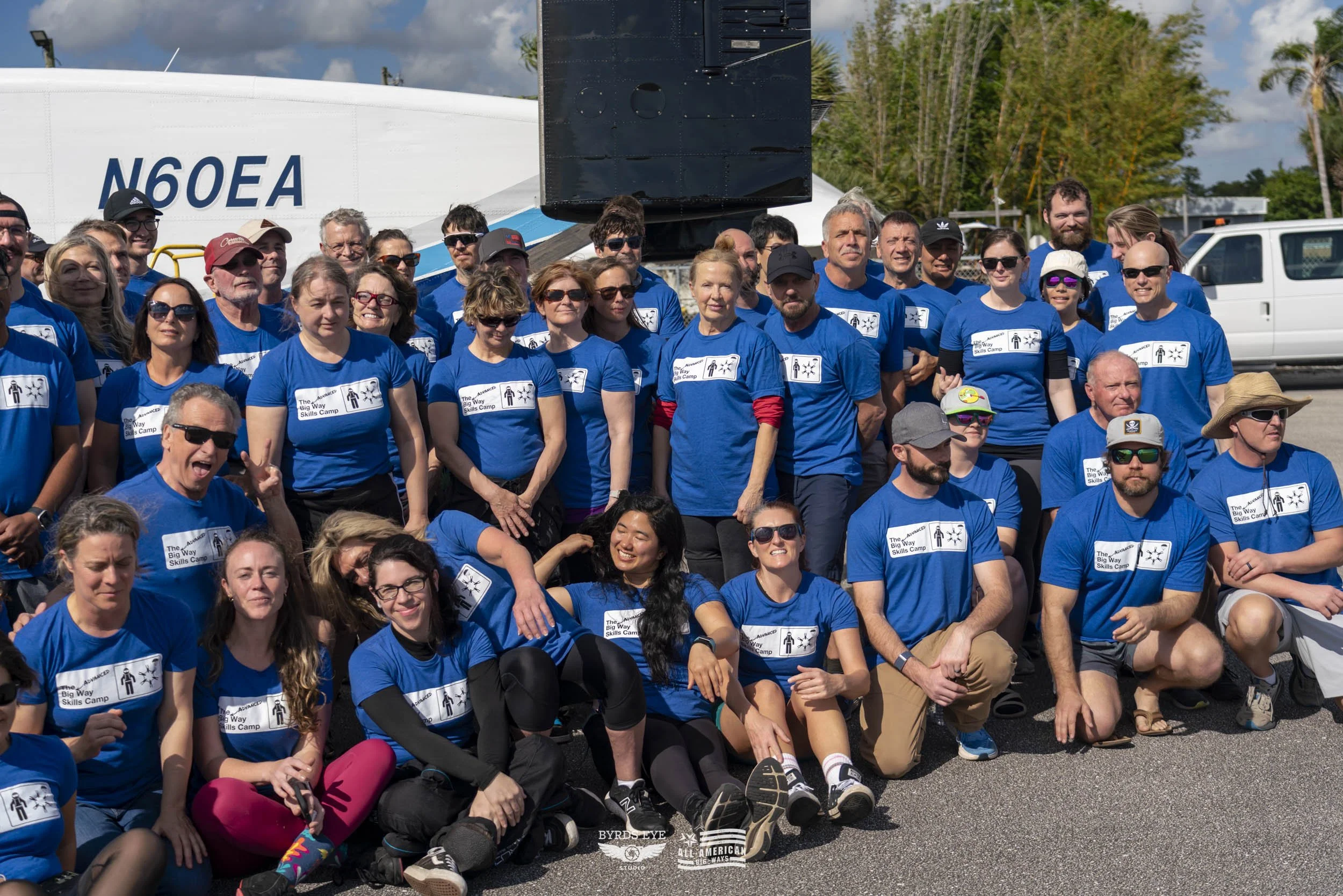Group of smiling people in blue T-shirts gathered outdoors in front of a helicopter, posing for a photo on a sunny day.