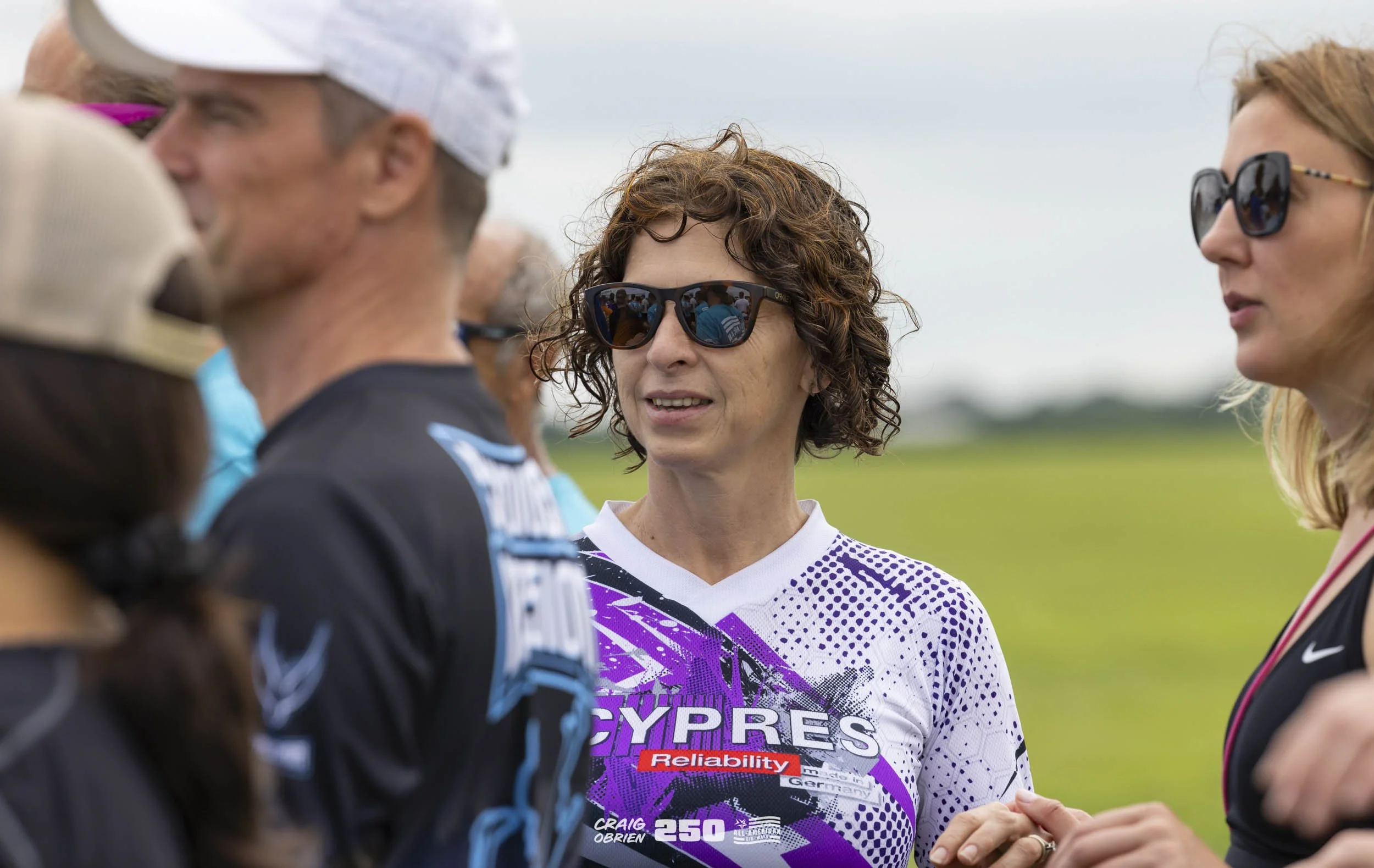 Group of people outdoors, woman in center wearing sunglasses and a cycling jersey talking to others during daytime.