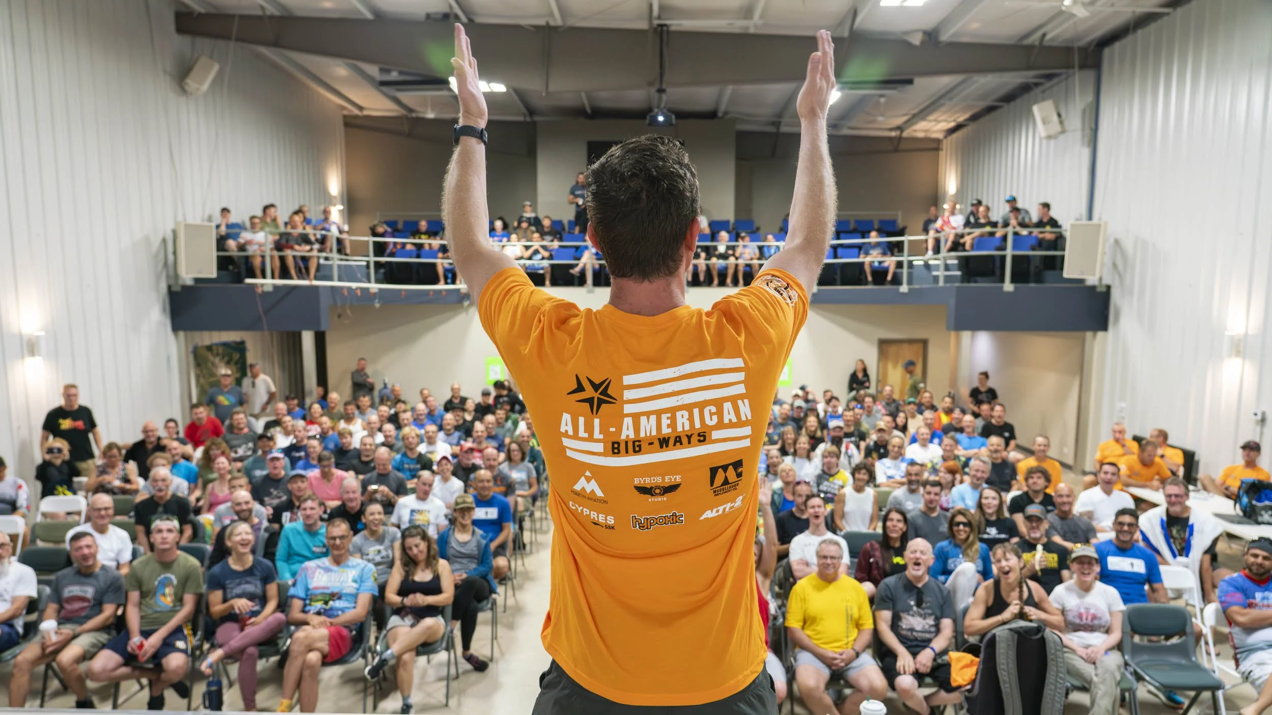 A man with short hair in an orange shirt stands on a stage with his arms raised, facing a large audience in a hall with a mezzanine. The audience members are seated and appear to be engaged and amused. The shirt has the words "ALL-AMERICAN BIG-WAYS" along with various logos.
