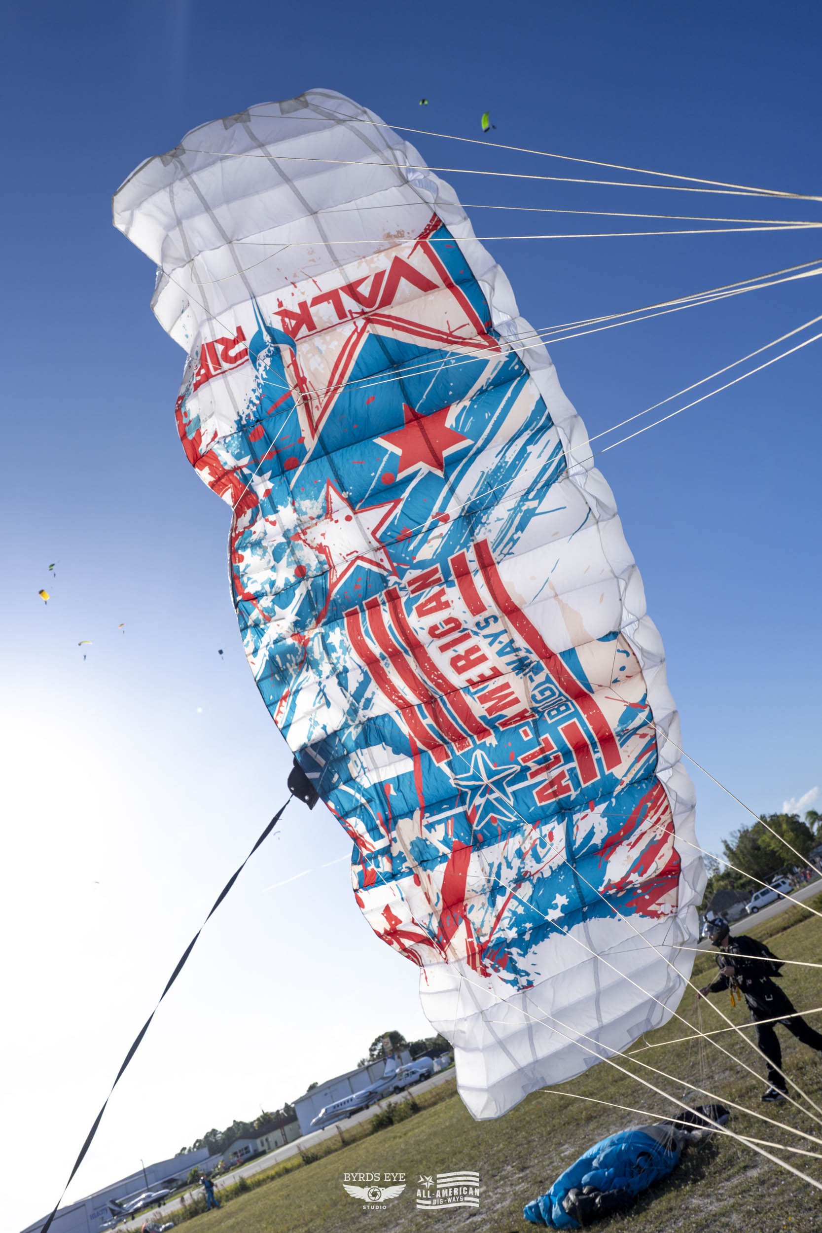 A large parachute with red, blue, and white patterns and text, flying in the sky during a sunny day, with other smaller parachutes visible in the background.