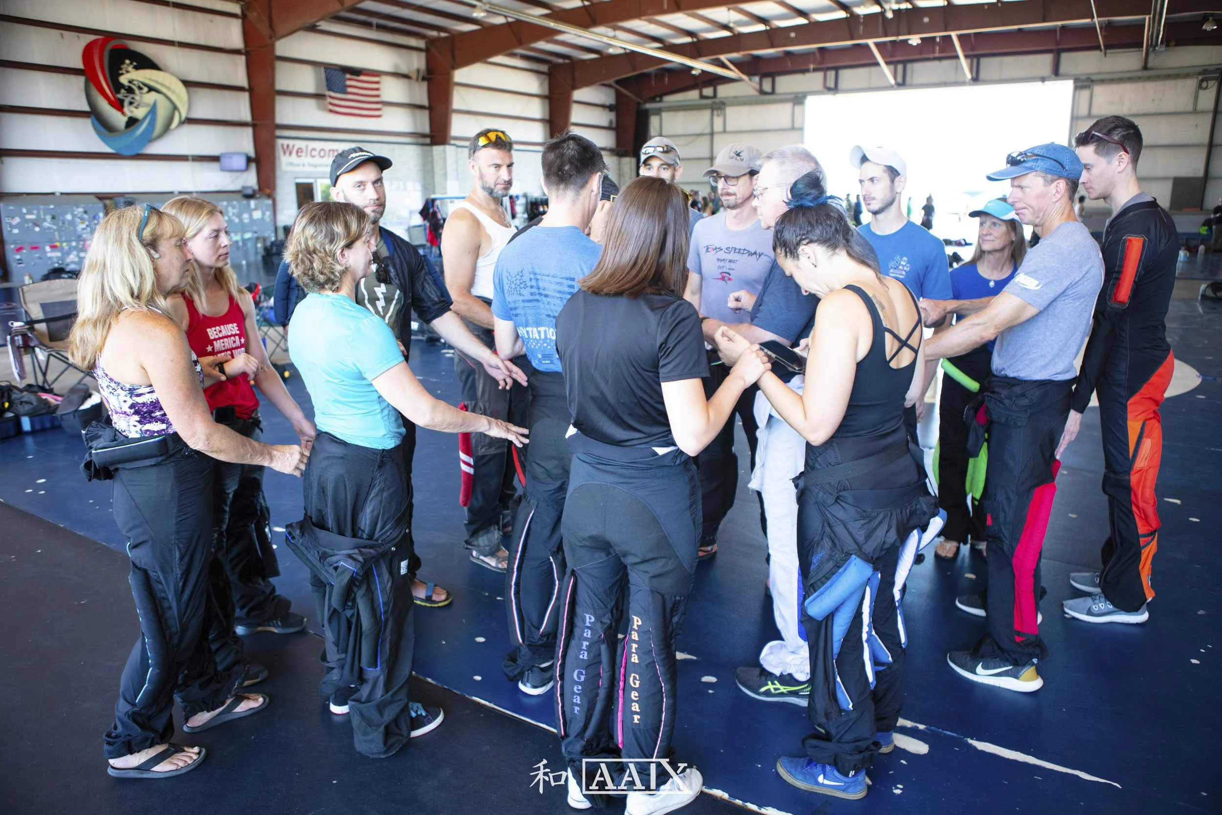 Group of skydivers gathering inside a hangar, some wearing jumpsuits, with a welcome sign and American flag in the background.