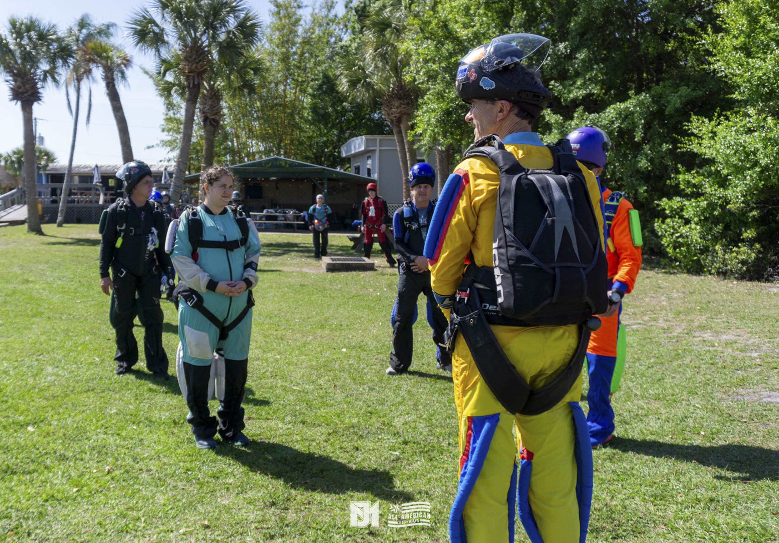 A group of skydivers gather outdoors on a grassy field, wearing jumpsuits and helmets, with trees and a pavilion in the background.