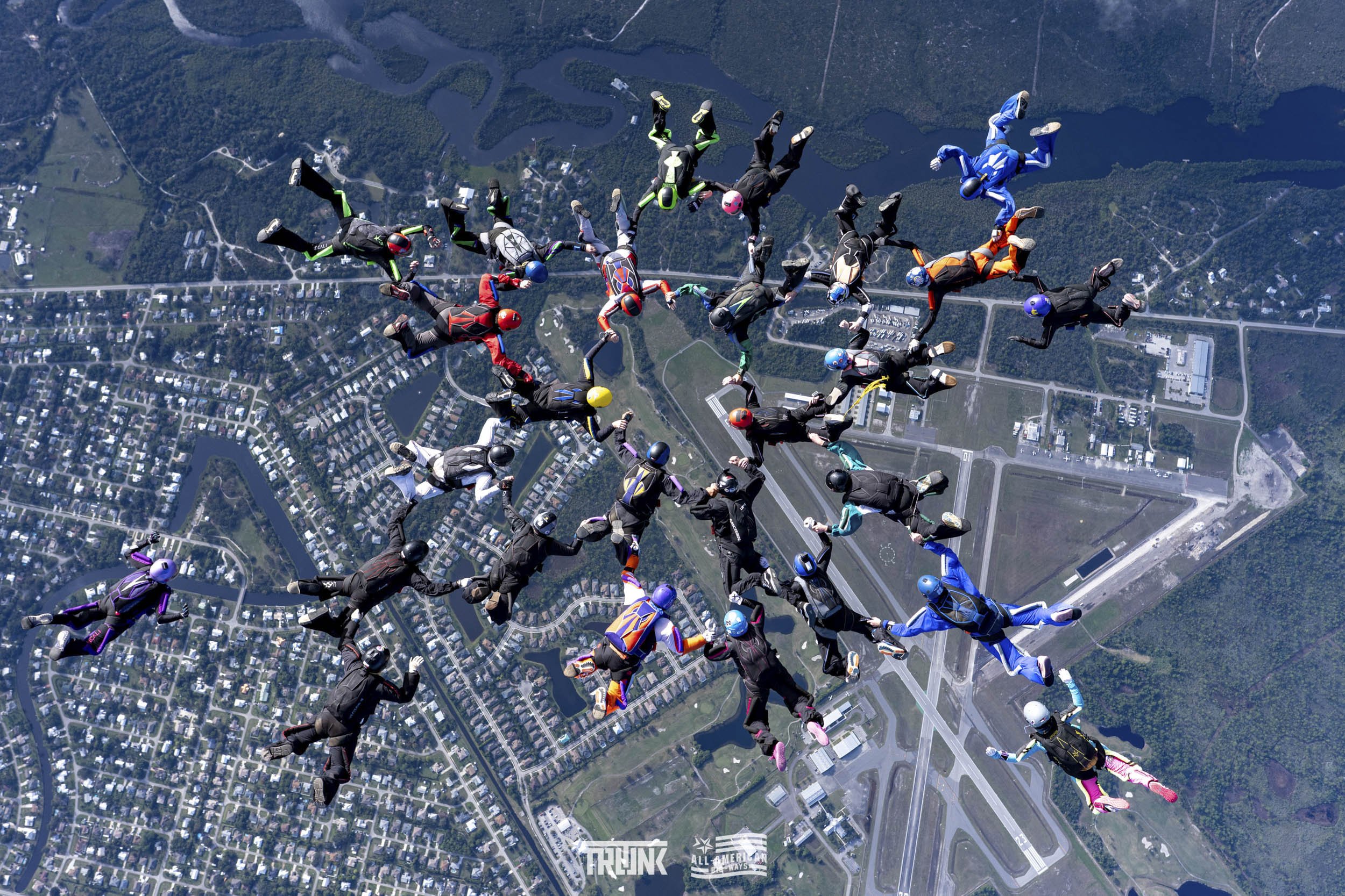 Group of skydivers forming a circle during a skydiving jump over a town with roads, bridges, and green areas.