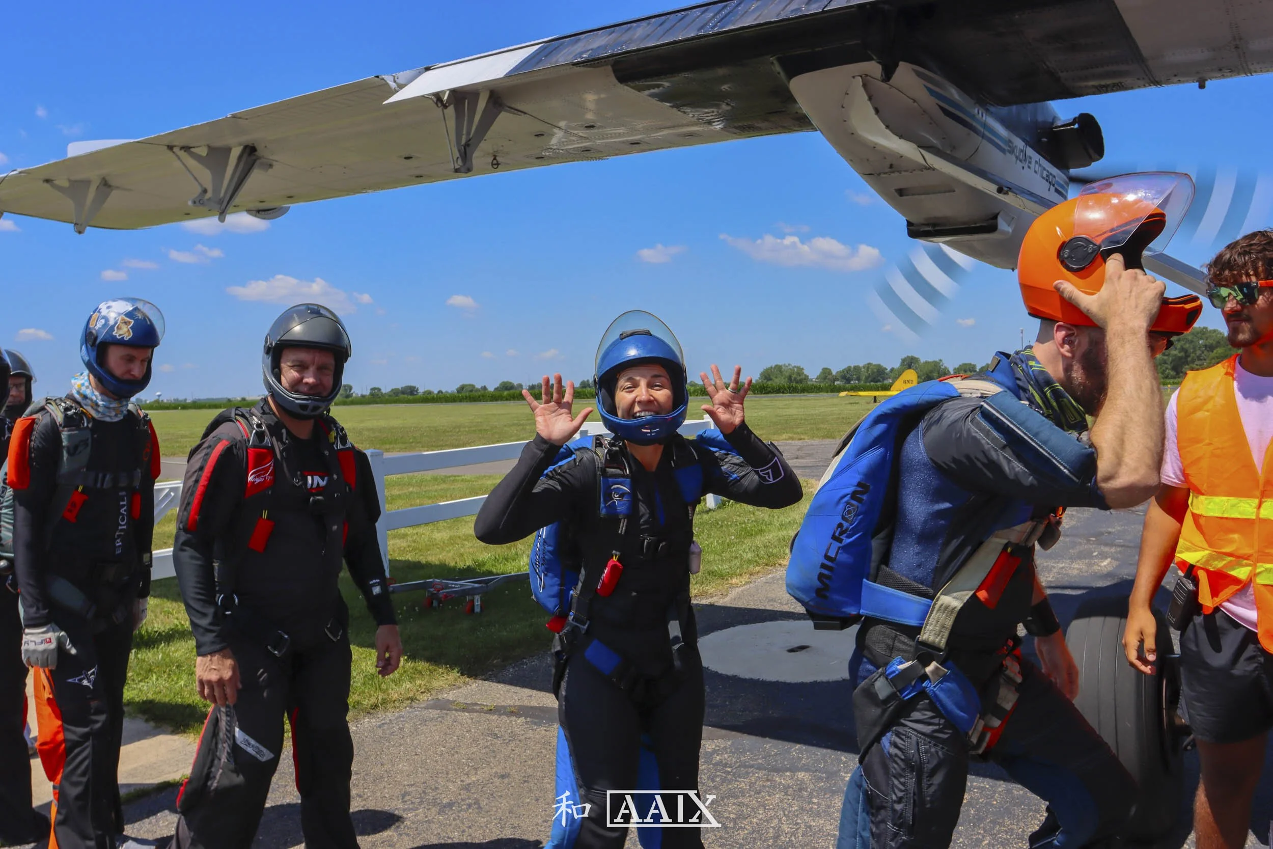 Group of skydivers with parachute gear next to an airplane on a grassy airfield under a blue sky, one smiling woman making a peace sign, others preparing for skydiving.