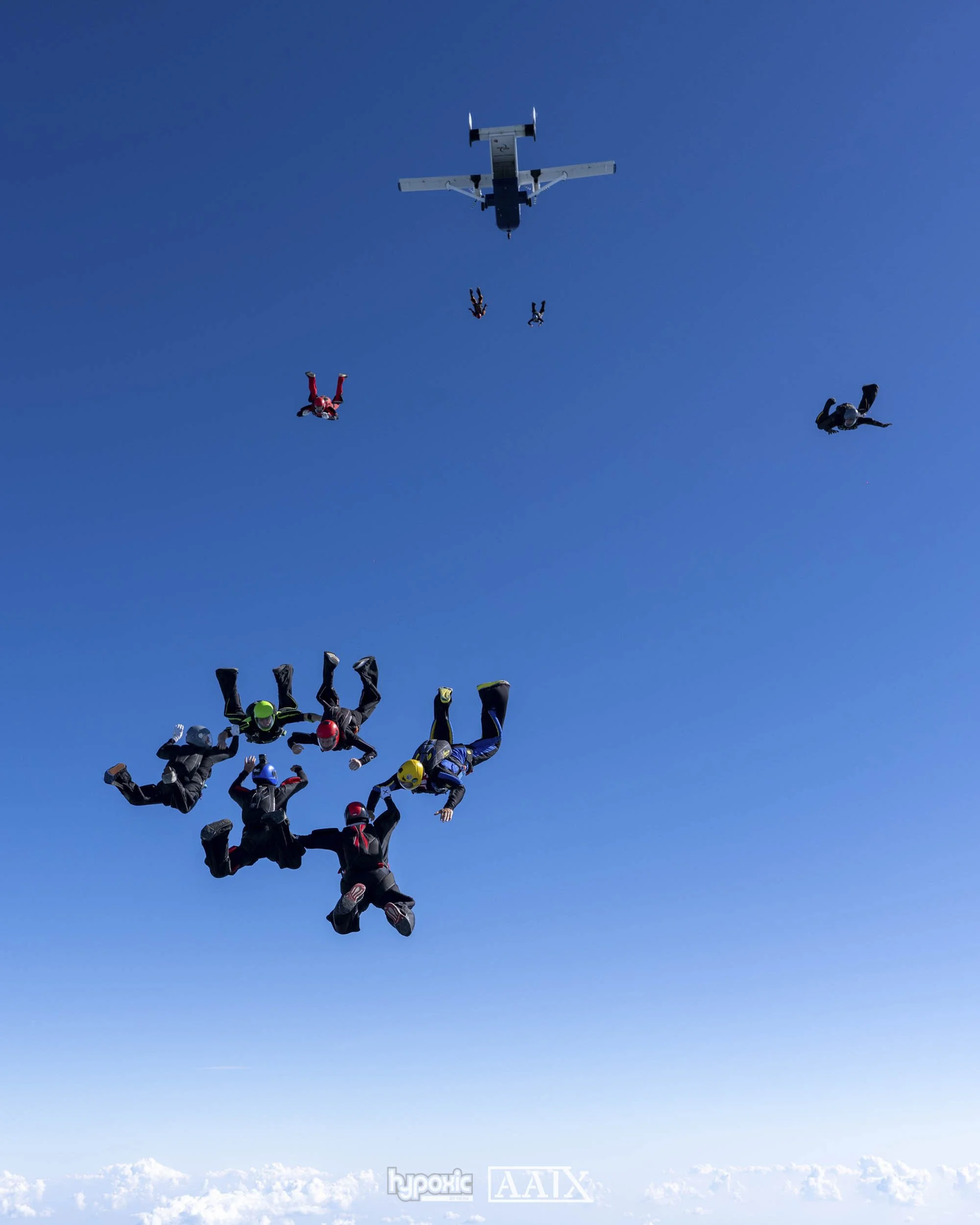 Skydivers in free fall formation with a small airplane overhead on a clear blue sky.