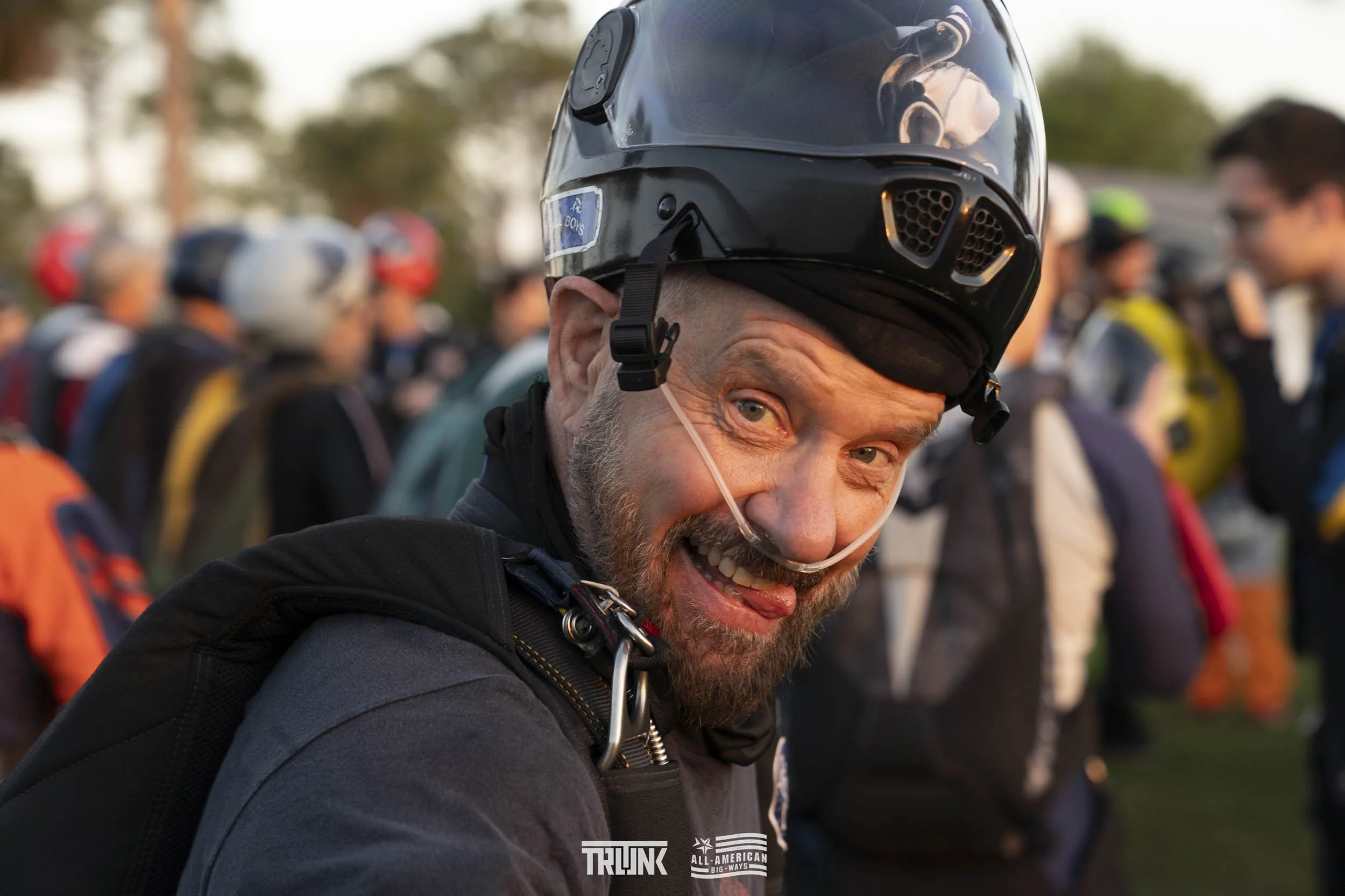 A smiling man wearing a motorcycle helmet and a nasal oxygen tube, surrounded by a group of people outdoors during daytime.