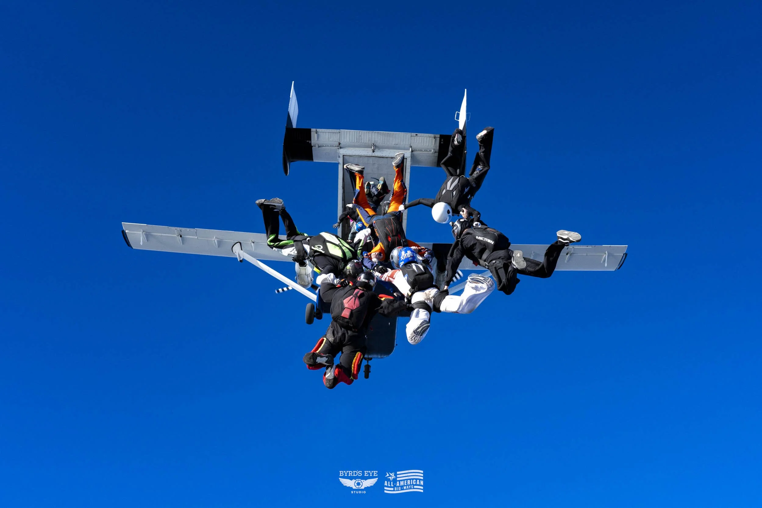 Skydivers holding onto an aircraft in freefall against a bright blue sky.