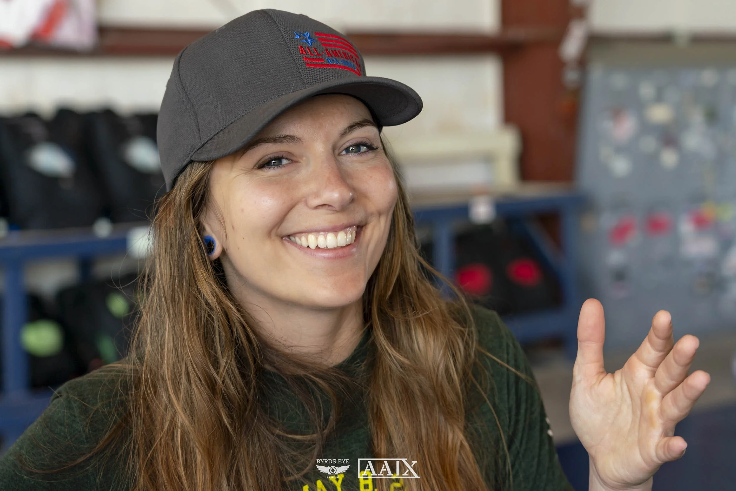 A smiling woman with long red hair wearing a gray cap and black shirt, in an indoor setting with shelves and equipment in the background.