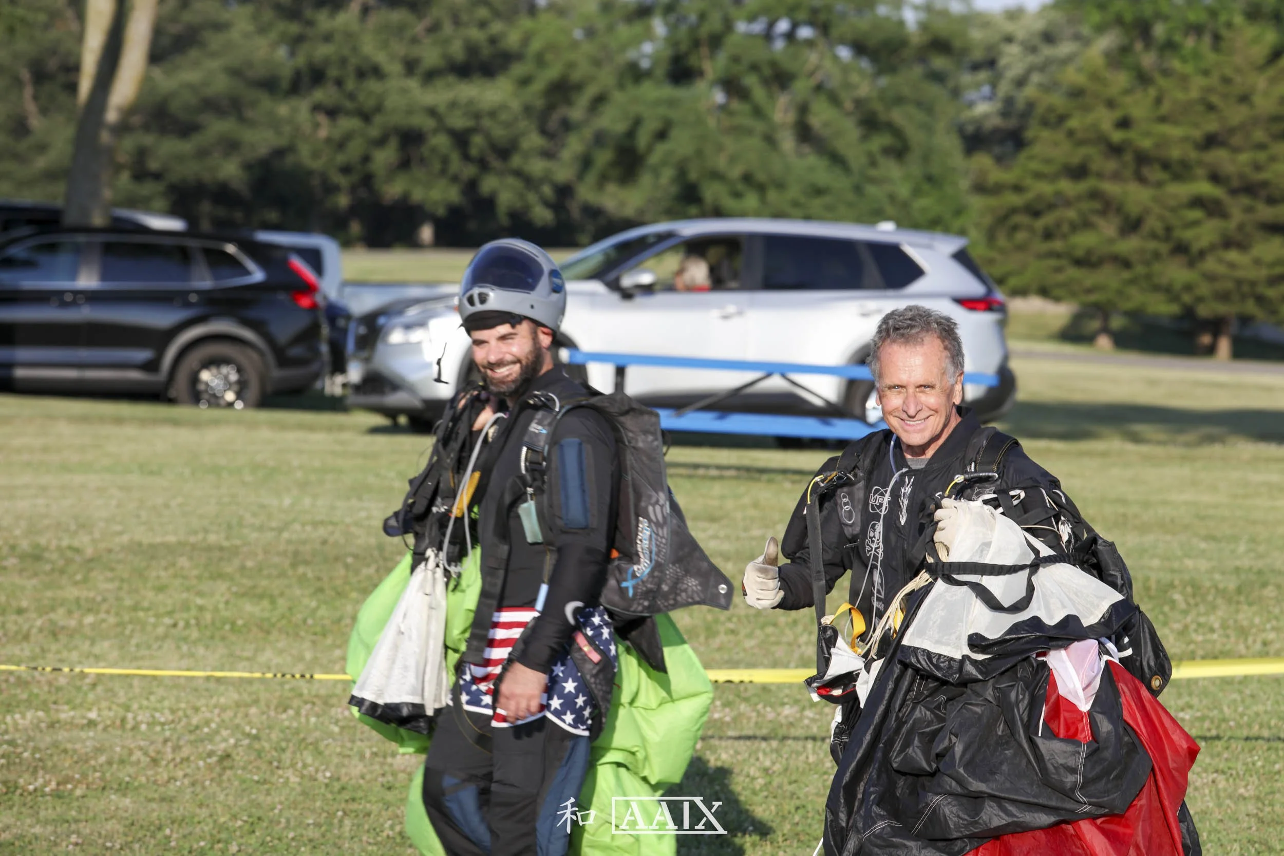 Two men in skydiving gear, smiling and walking after a jump, holding parachutes with cars and trees in the background.