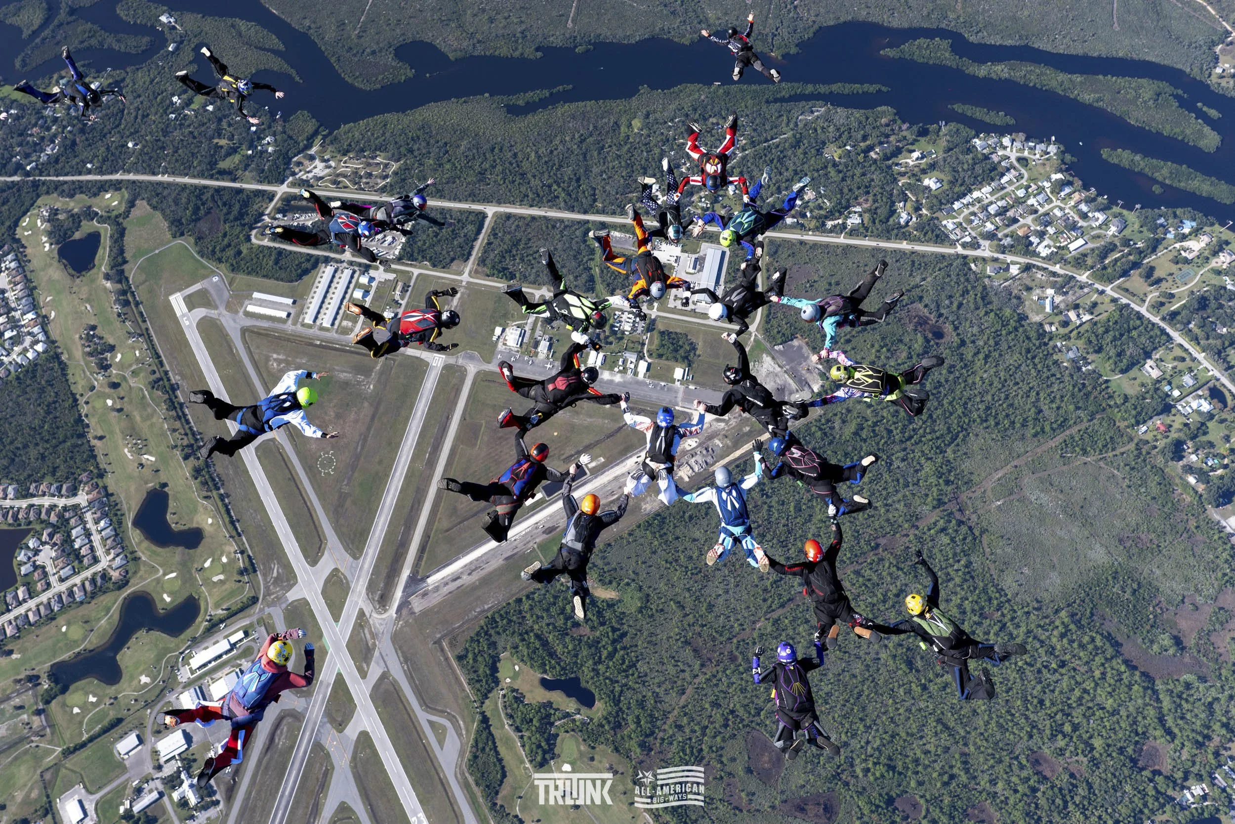 A large group of skydivers in colorful jumpsuits and helmets forming a circular pattern while free-falling over a patchwork of green land, roads, and small lakes.