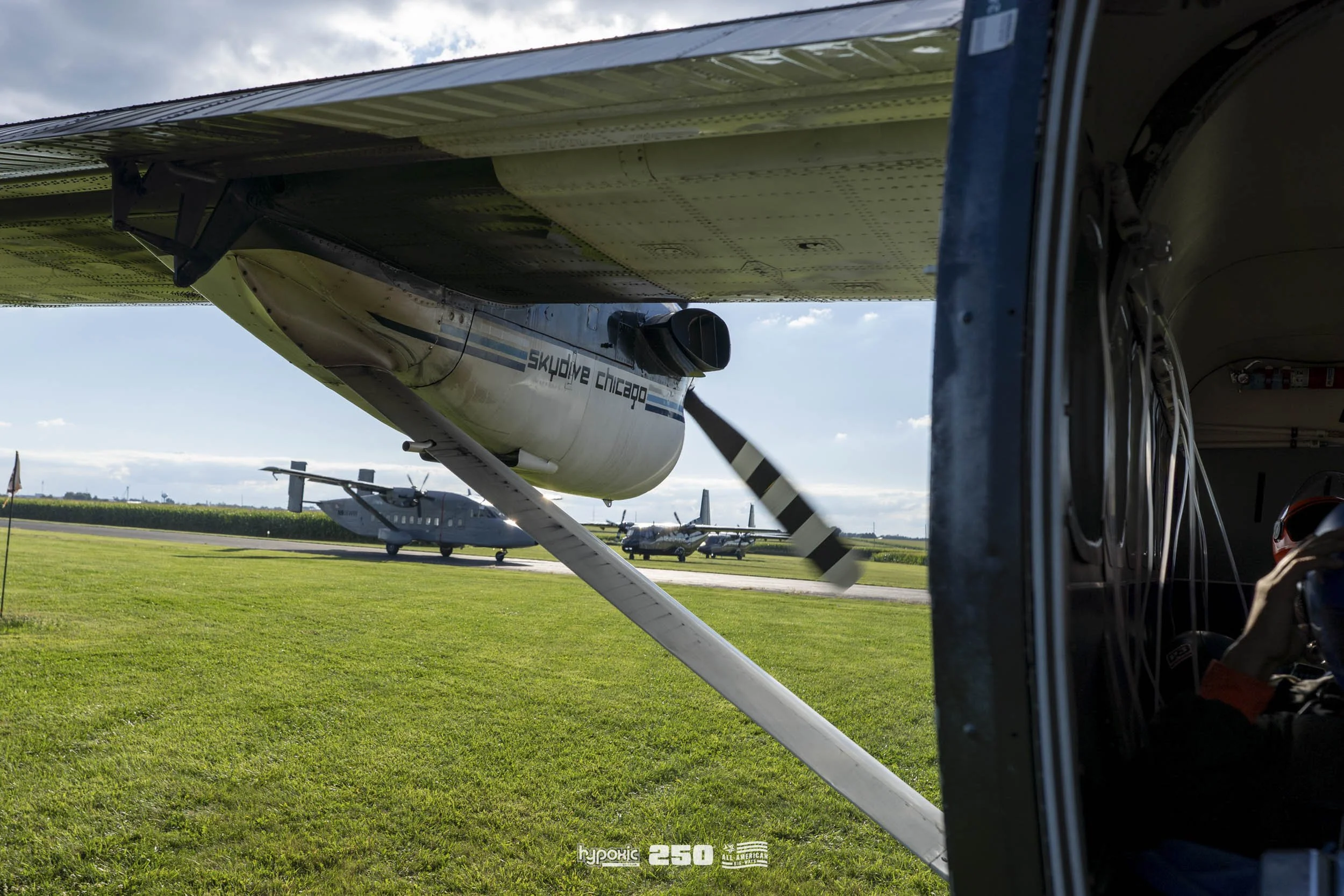 Close-up of a small airplane wing with the text 'skydive chicago' on it. In the background, there are two large aircraft on the ground at an airport, with a grassy field and blue sky with some clouds.