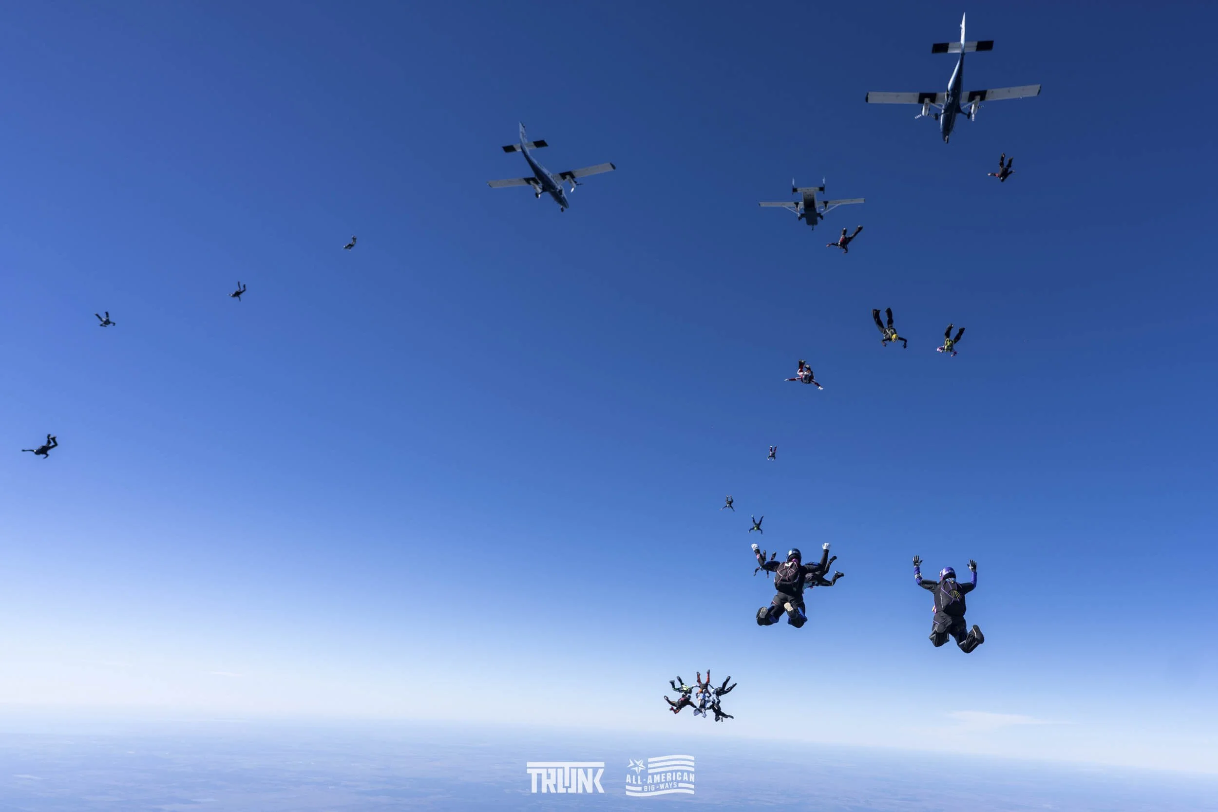 Skydivers free-falling with airplanes in the background against a clear blue sky.