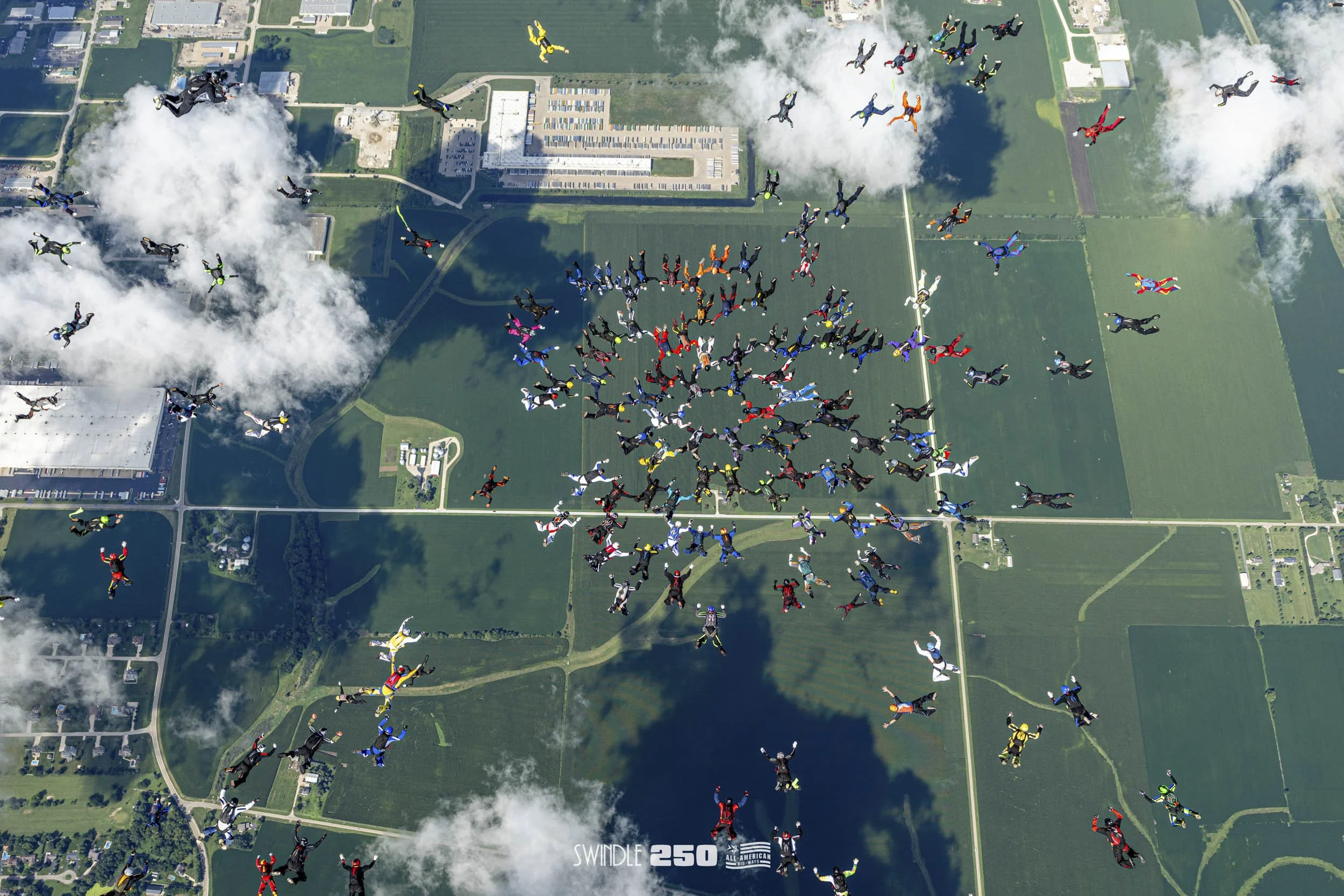 A large group of skydivers in colorful jumpsuits are skydiving over a landscape with green fields, residential areas, and industrial buildings. The skydivers are spread across the sky, with some near clouds and others closer to the ground, casting a 