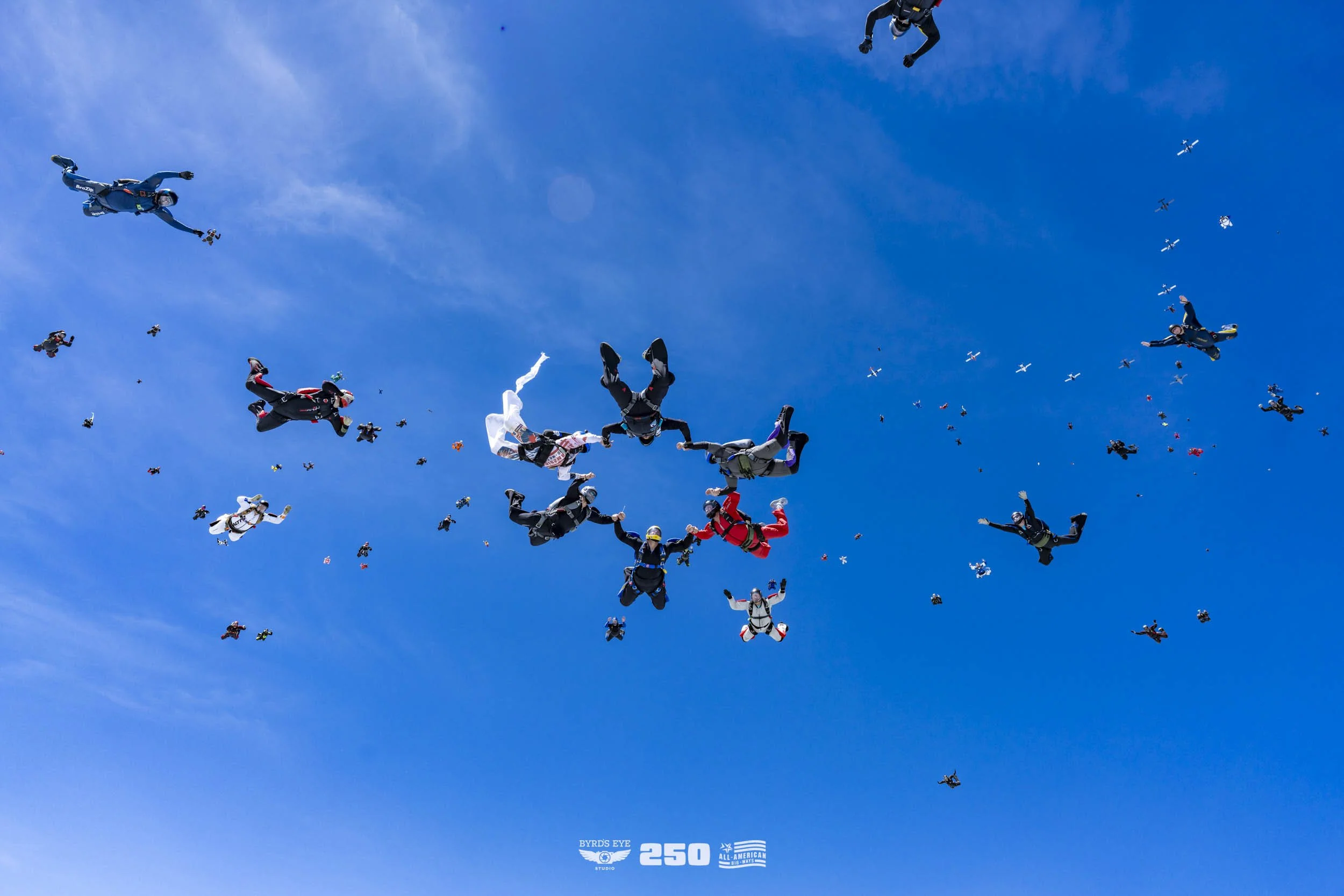 A large group of skydivers in colorful jumpsuits and parachutes free-falling against a bright blue sky with some small clouds.