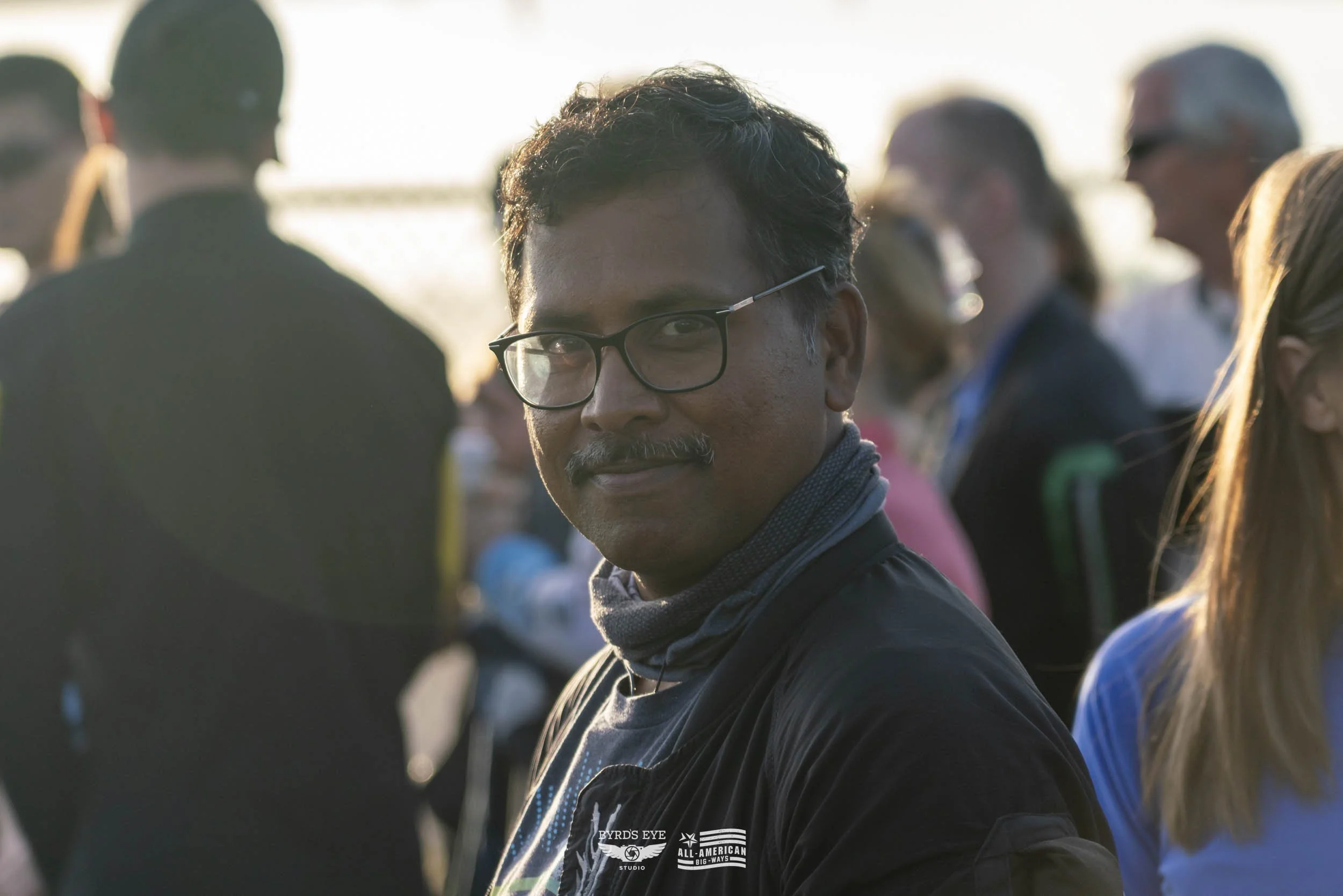 A man with glasses and a mustache smiling at the camera during an outdoor event with a crowd in the background.
