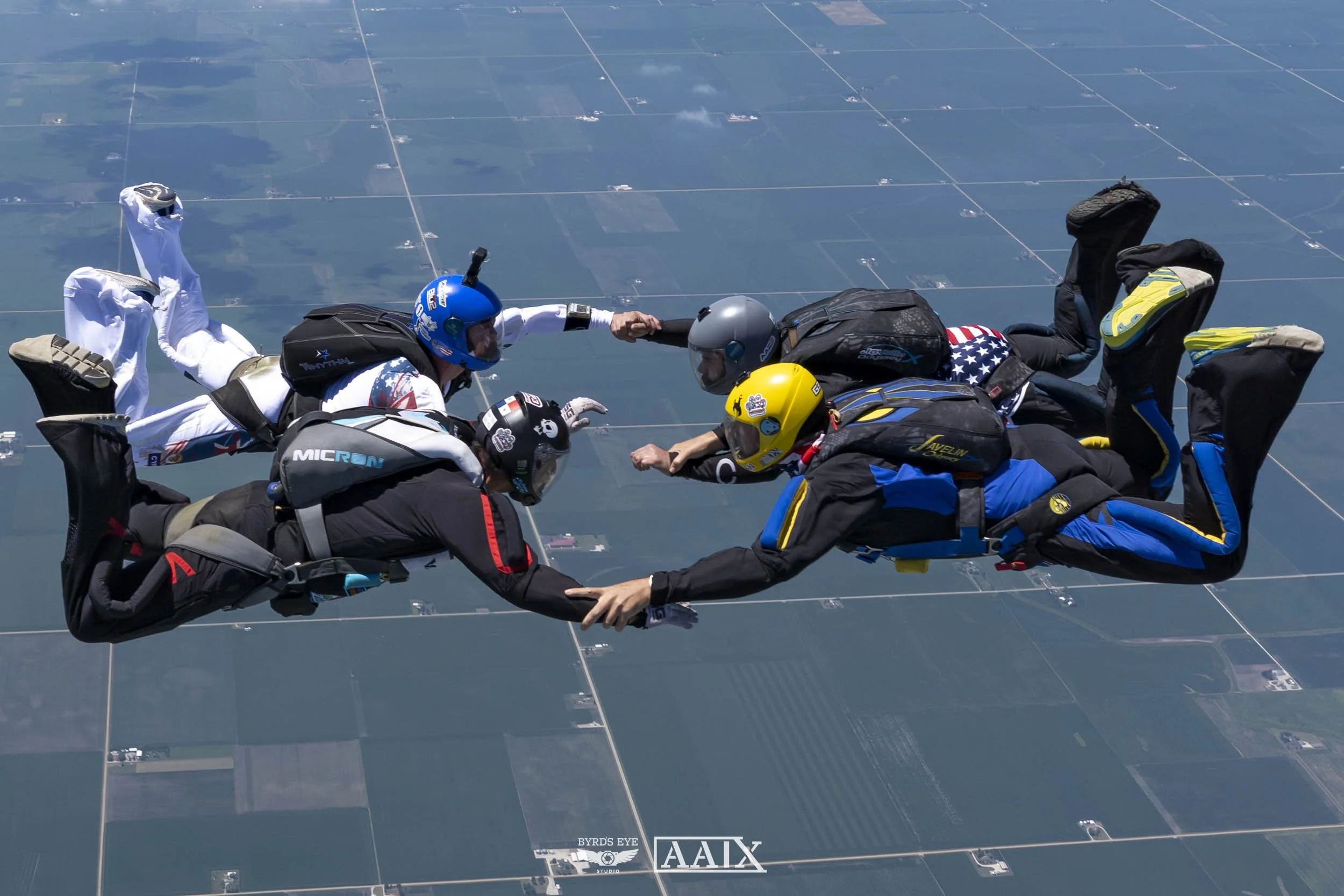 Five skydivers holding hands in freefall over farmland, wearing helmets and jumpsuits.