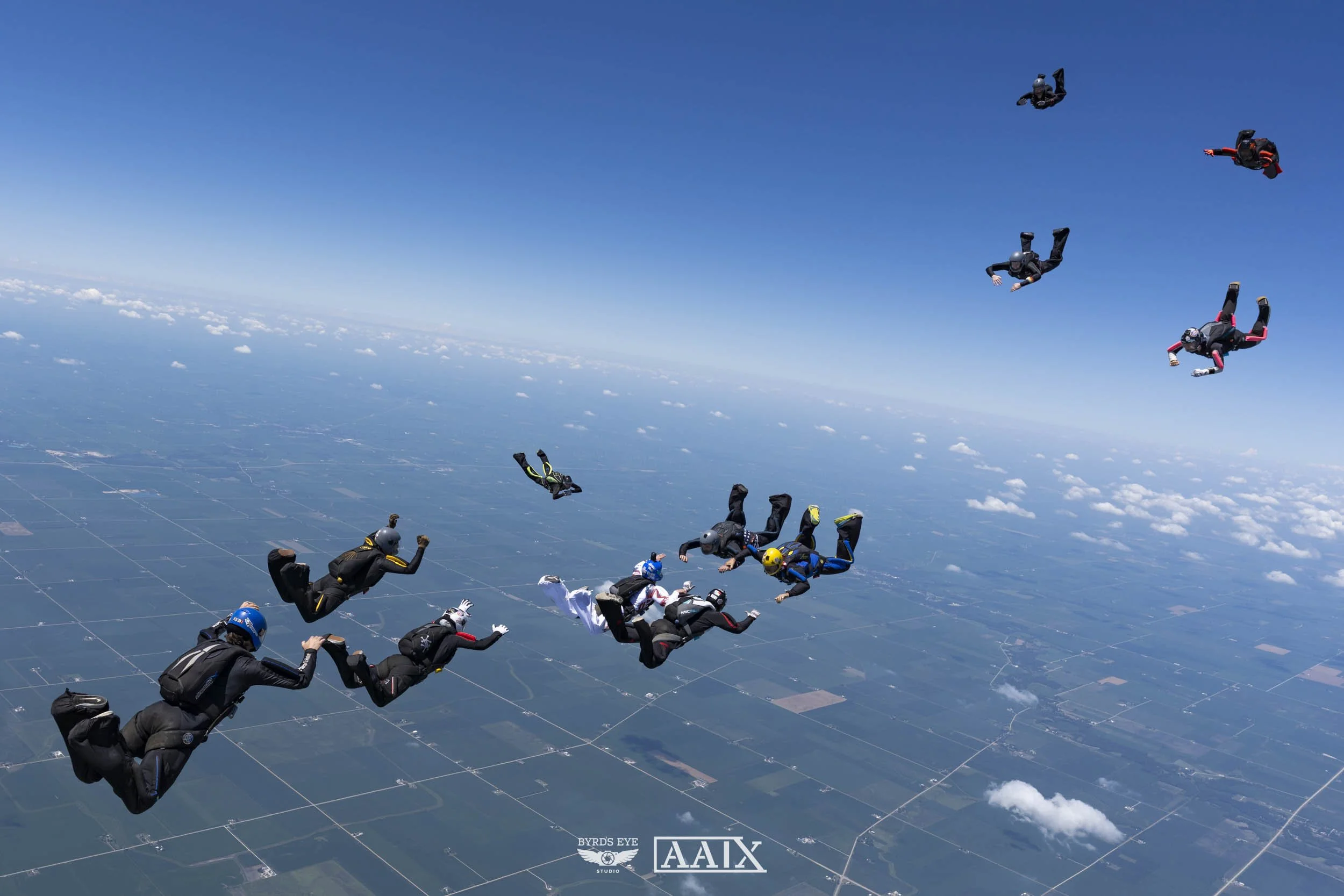 Group of skydivers free-falling through the sky over a landscape with clouds and fields below.