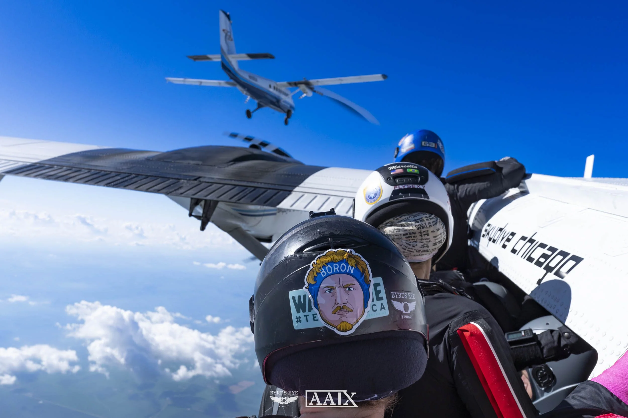 Skydivers in the aircraft ride, with one holding onto the wing, in mid-air, looking at the airplane flying above cloudy sky.