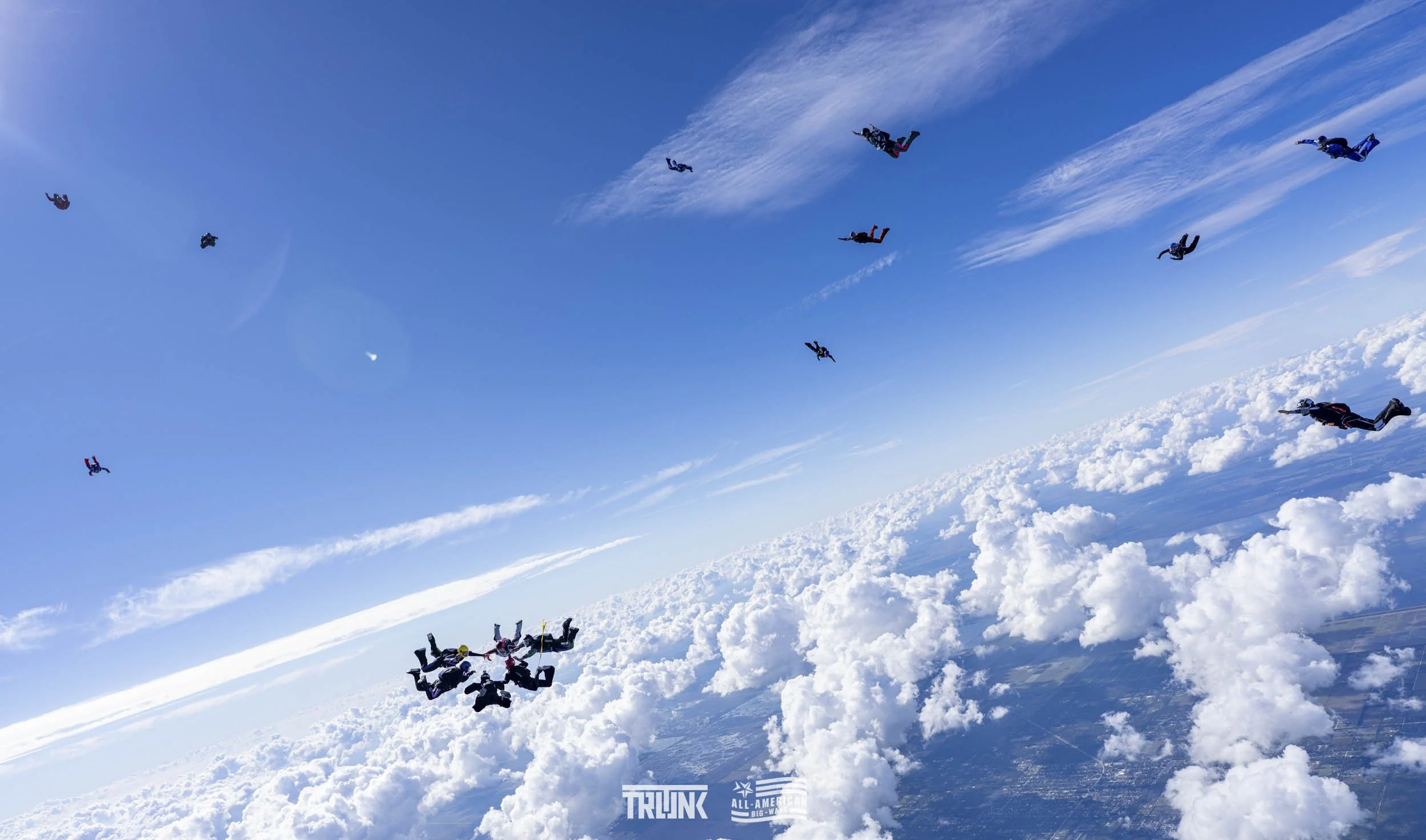 Skydivers in free fall over a cloud-covered landscape with blue sky and scattered clouds.