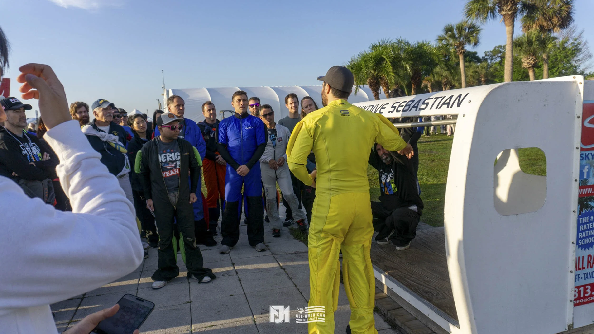 A group of skydivers gathered outdoors in front of a small aircraft, listening to a man in a yellow jumpsuit. Palm trees and a clear blue sky are in the background.