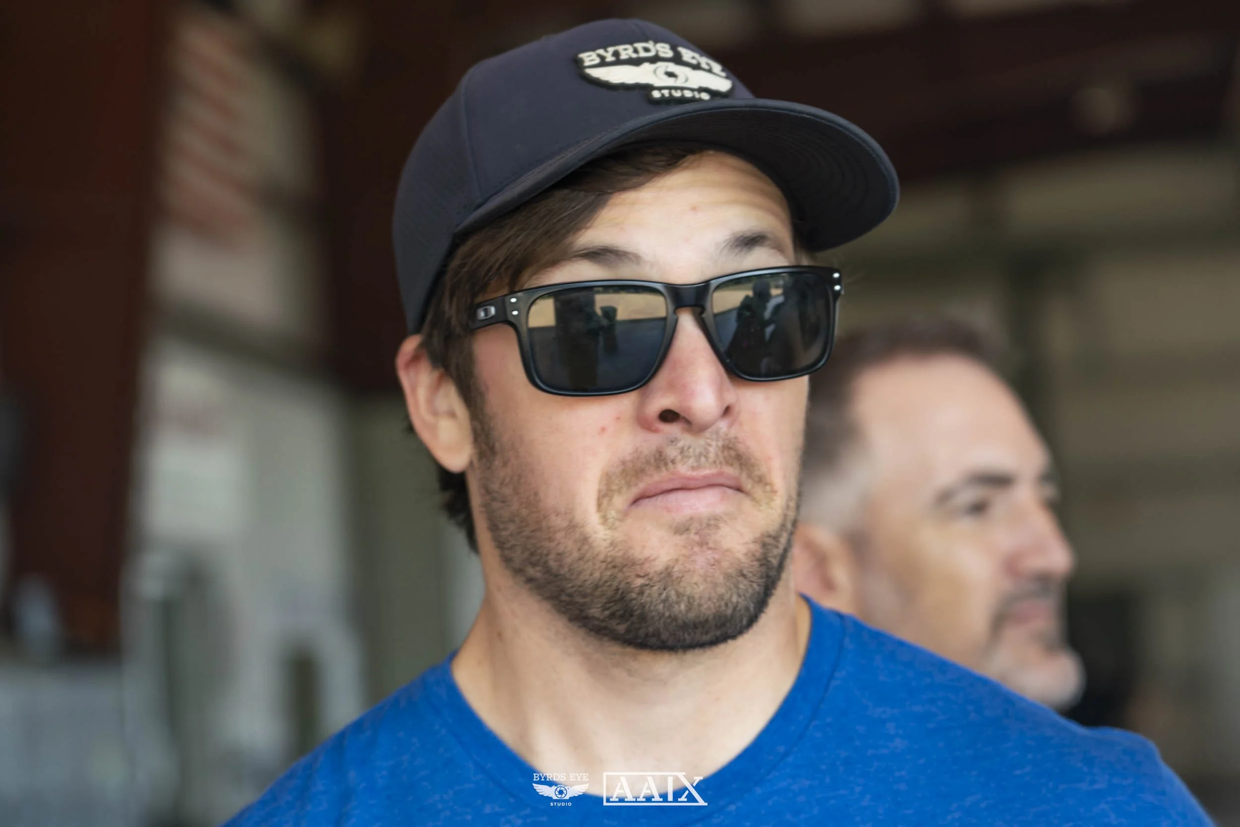 A man wearing large black sunglasses, a blue t-shirt, and a black cap with a Byrd's Eye logo, standing indoors with another man blurred in the background.