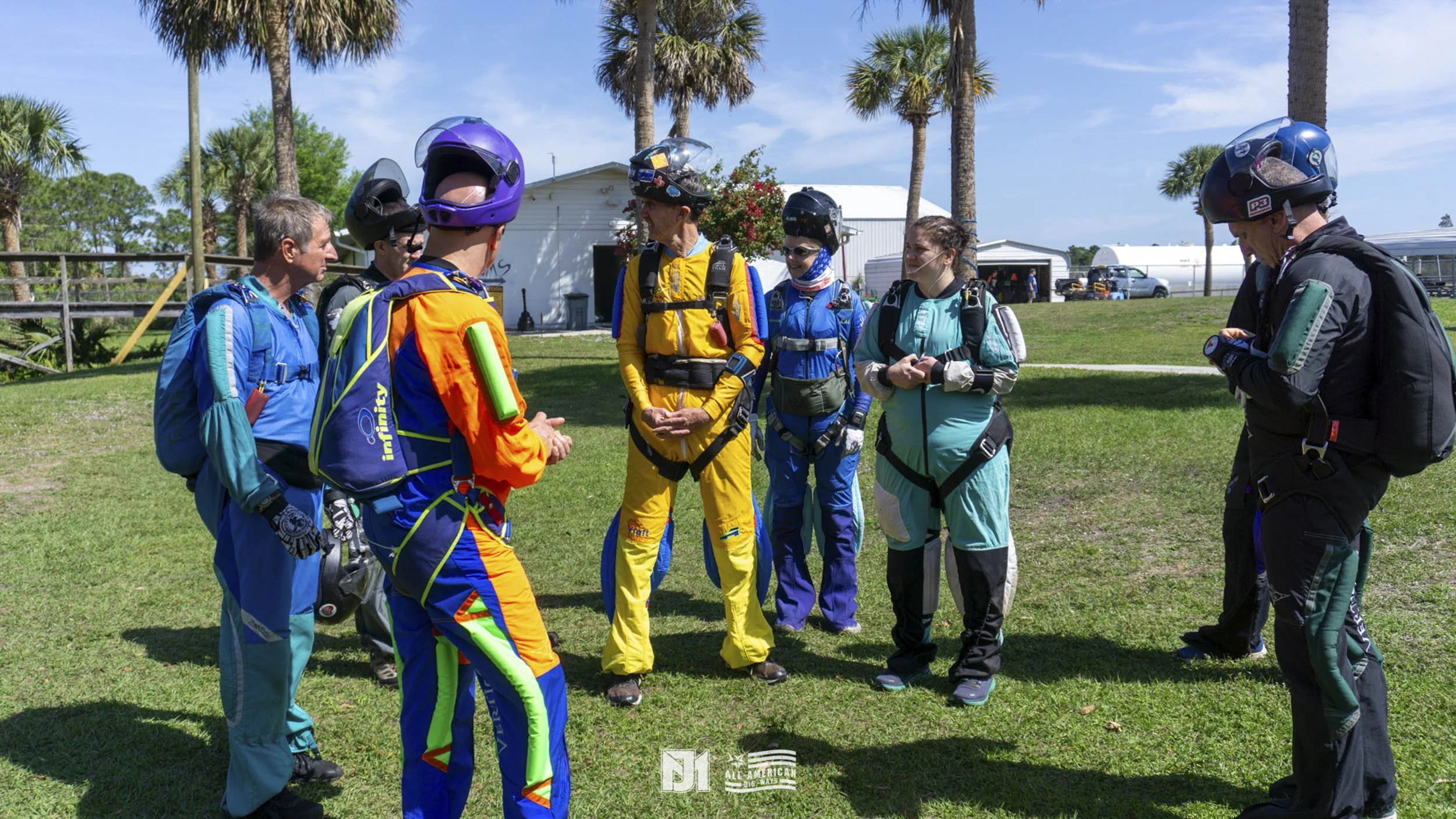 A group of skydivers wearing colorful jumpsuits and helmets standing on grass, listening to an instructor during briefing outdoors with palm trees and a clear blue sky.