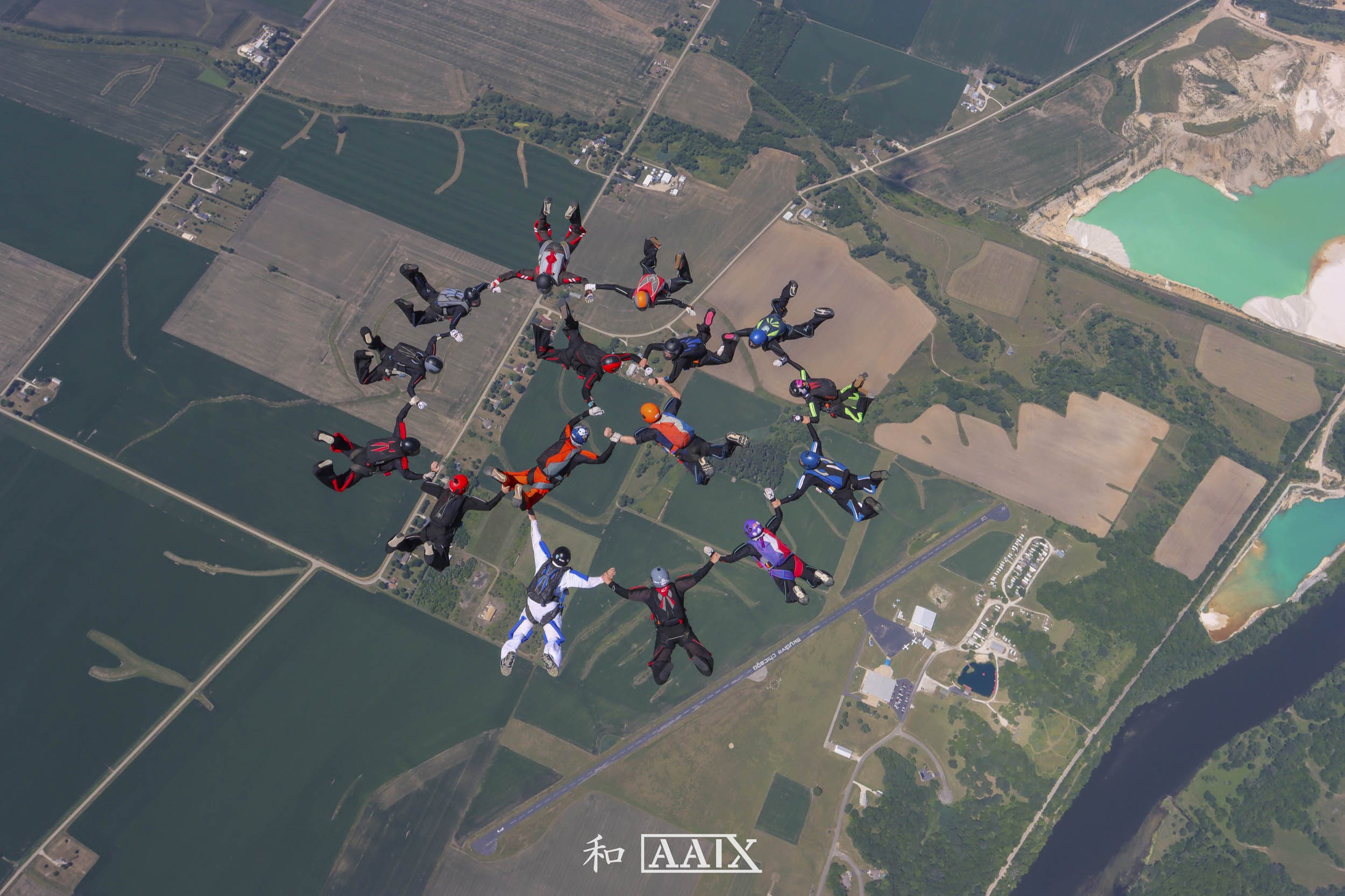 Group of skydivers in jumpsuits and helmets forming a circle while free-falling over a landscape with fields, a body of water, and a lake.