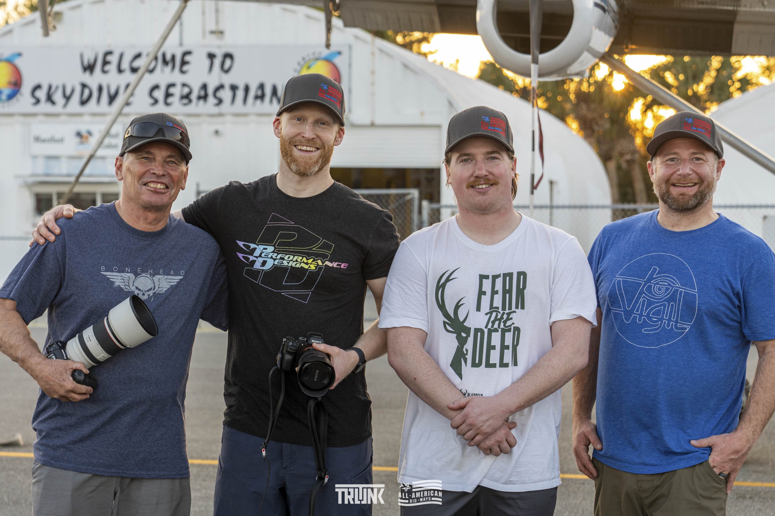 Four men standing together outdoors at sunset, smiling at the camera, with a skydiving center in the background that reads 'Welcome to Skydiving Sebastian.' They are casual dressed and wearing hats, two holding cameras, and are standing on a tarmac w