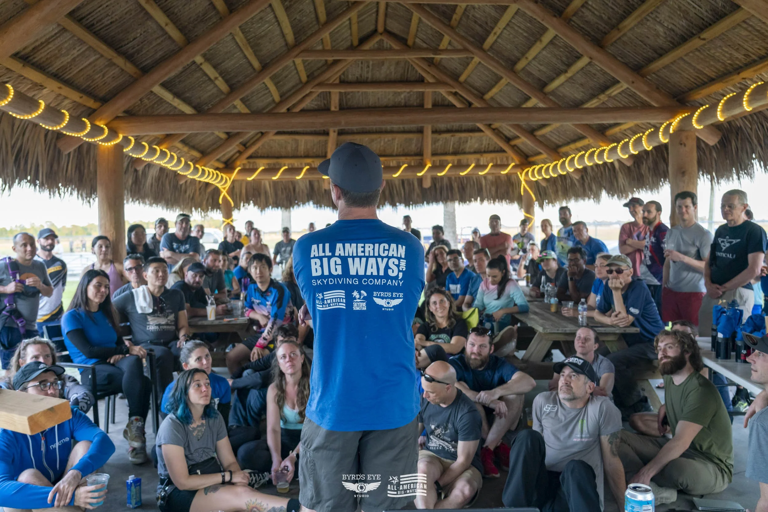 A person in a blue shirt with white text speaking to a large group seated under a thatched roof pavilion at an outdoor event.