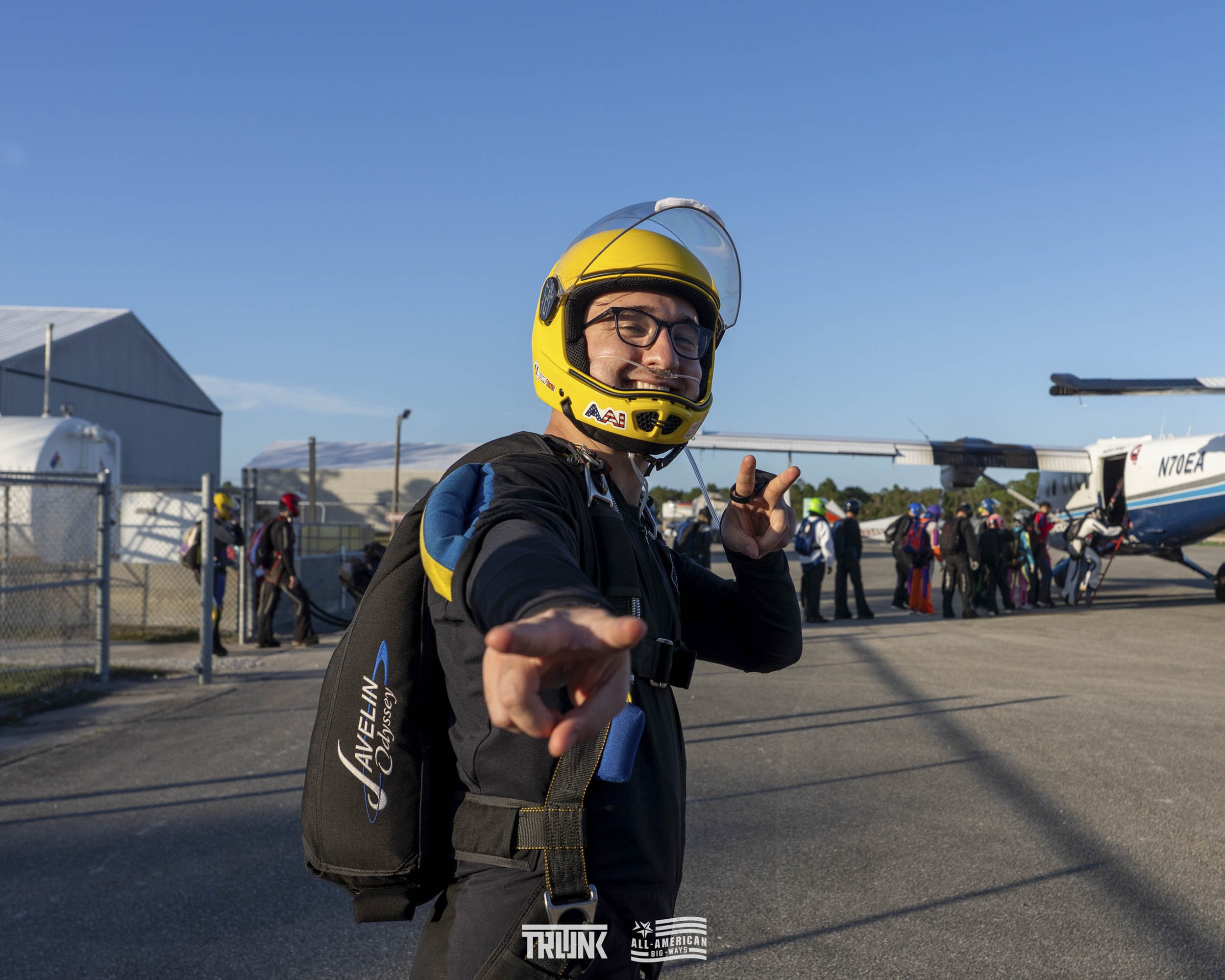 Person in a motorcycle helmet and gear smiling and posing at the airport with a group of skydivers in the background.