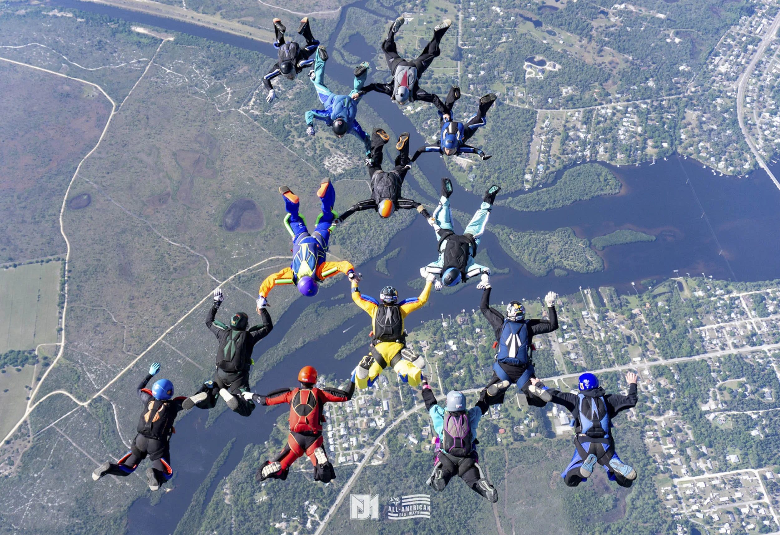A group of skydivers in colorful jumpsuits and helmets are forming a star shape while free-falling over a landscape with lakes, forests, and roads.