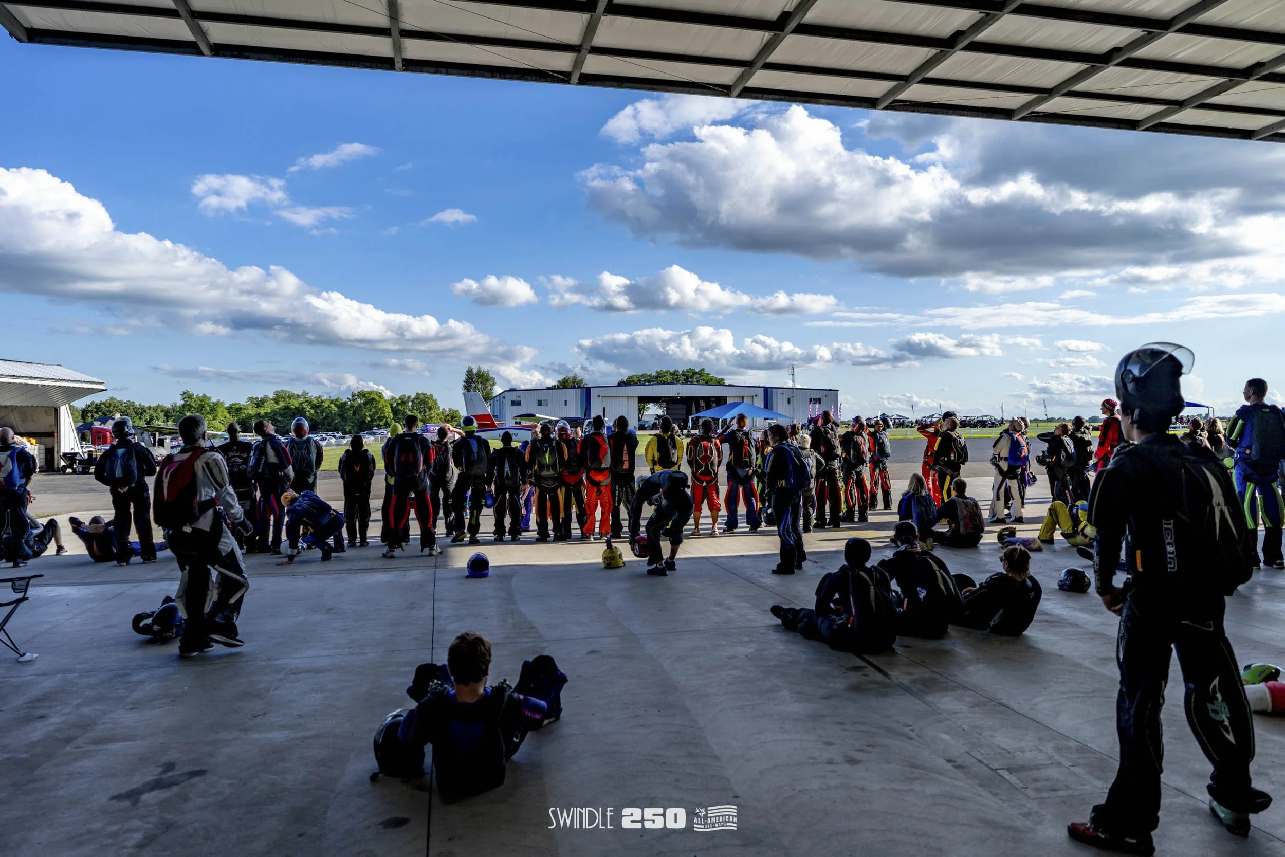 Skydivers gather at an airport with a large open hangar, clouds in the sky, and a small airplane on the tarmac.