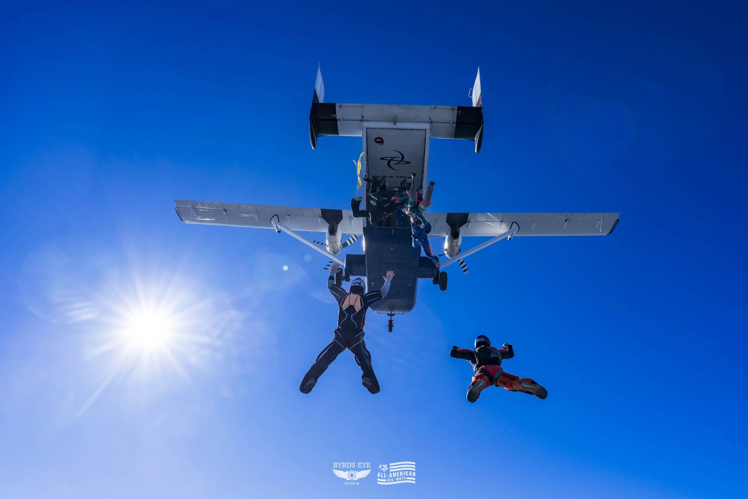 Skydivers exiting a small airplane against a clear blue sky with the sun shining brightly.