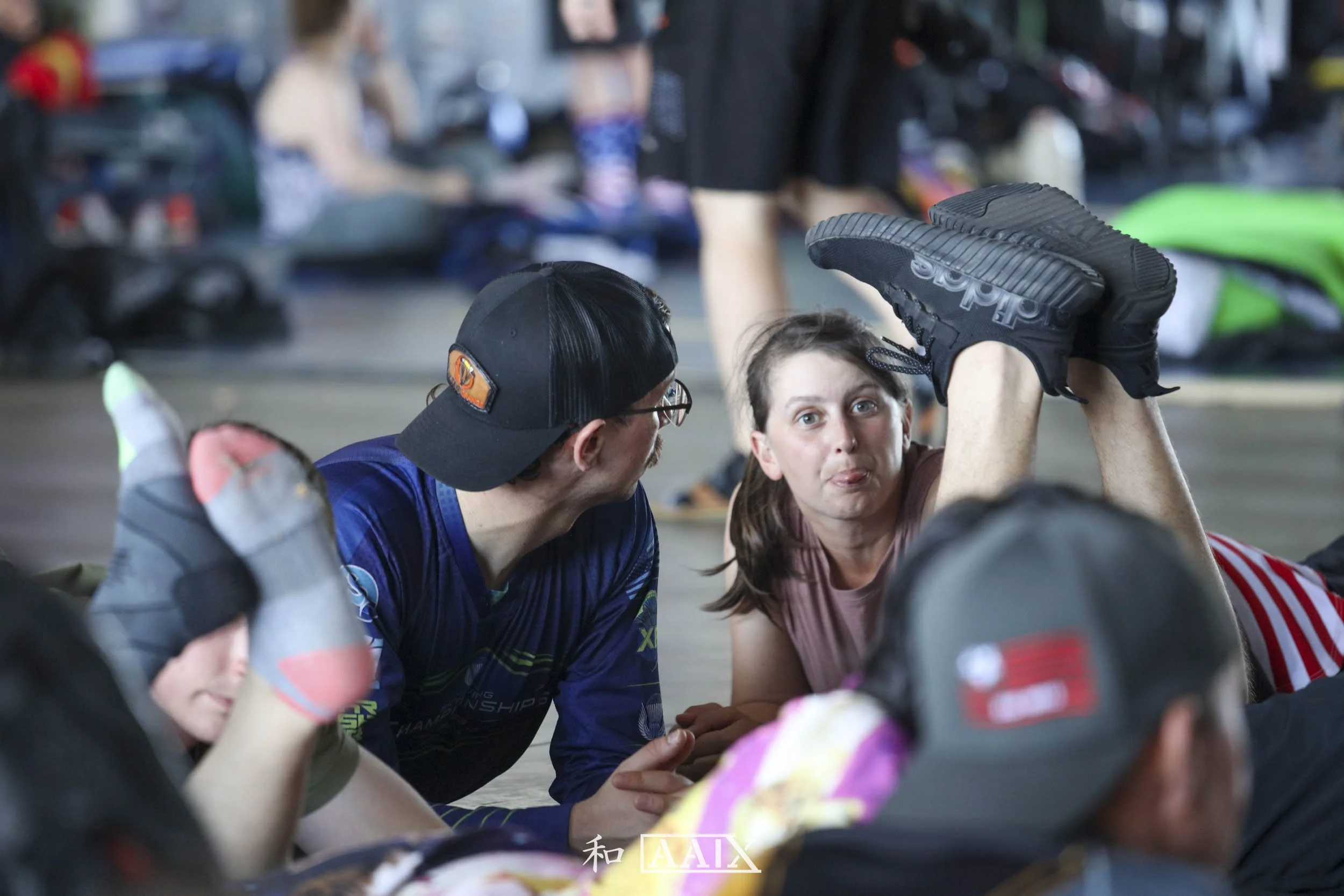 People lying on the floor, with one woman lifting her leg while wearing a sports shoe, and others looking at her, inside a busy indoor space.