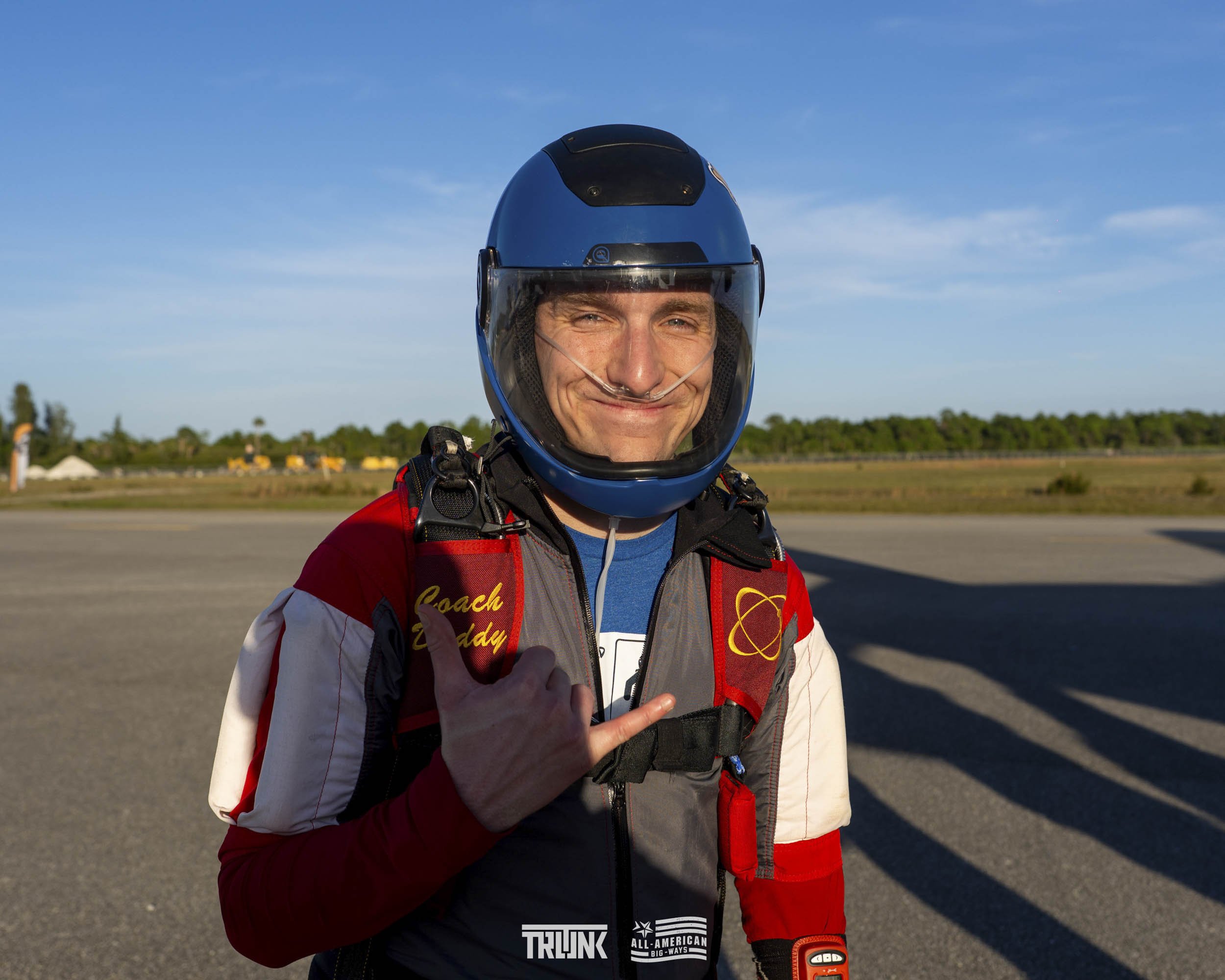 A smiling man wearing a blue helmet and a red racing suit, standing outdoors on a tarmac with a clear sky and distant trees, making a shaka sign with his right hand.
