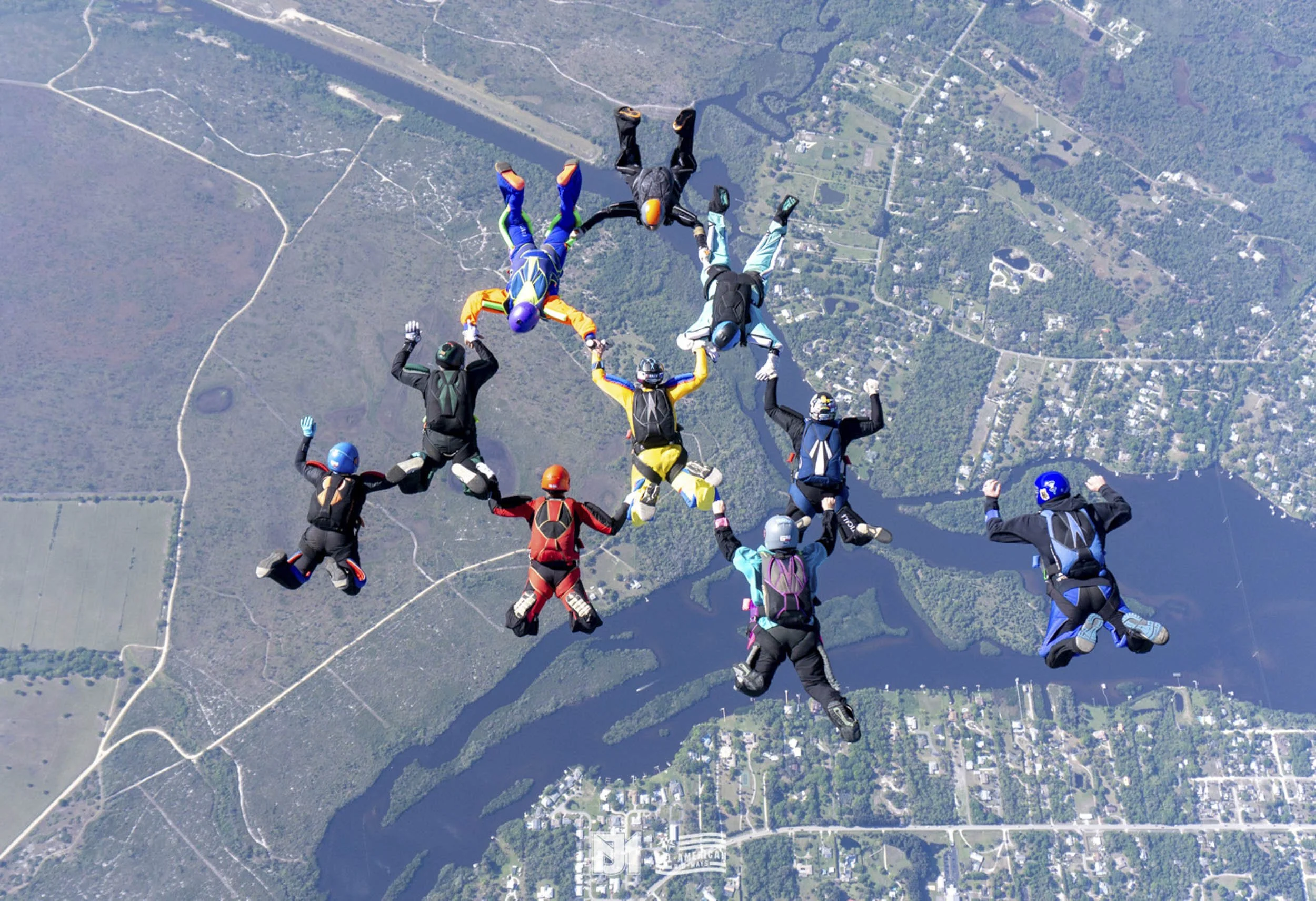 Group of skydivers holding hands mid-air over a landscape with water, trees, and roads.