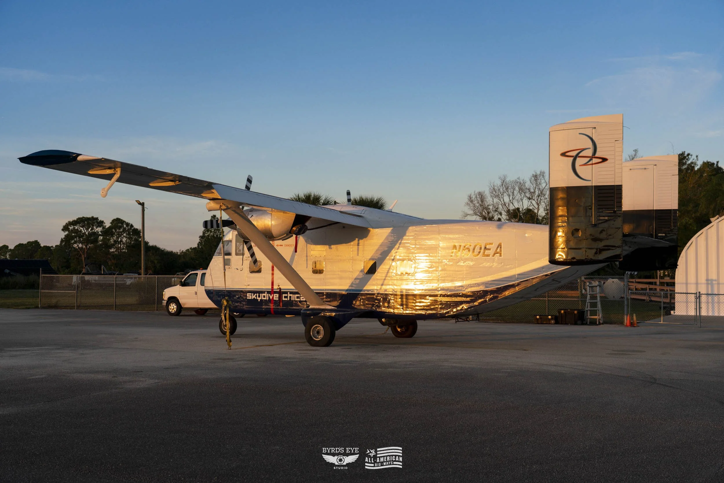 A small aircraft with a high wing design and twin engines parked on tarmac at sunset, with a vehicle and storage hangars in the background.